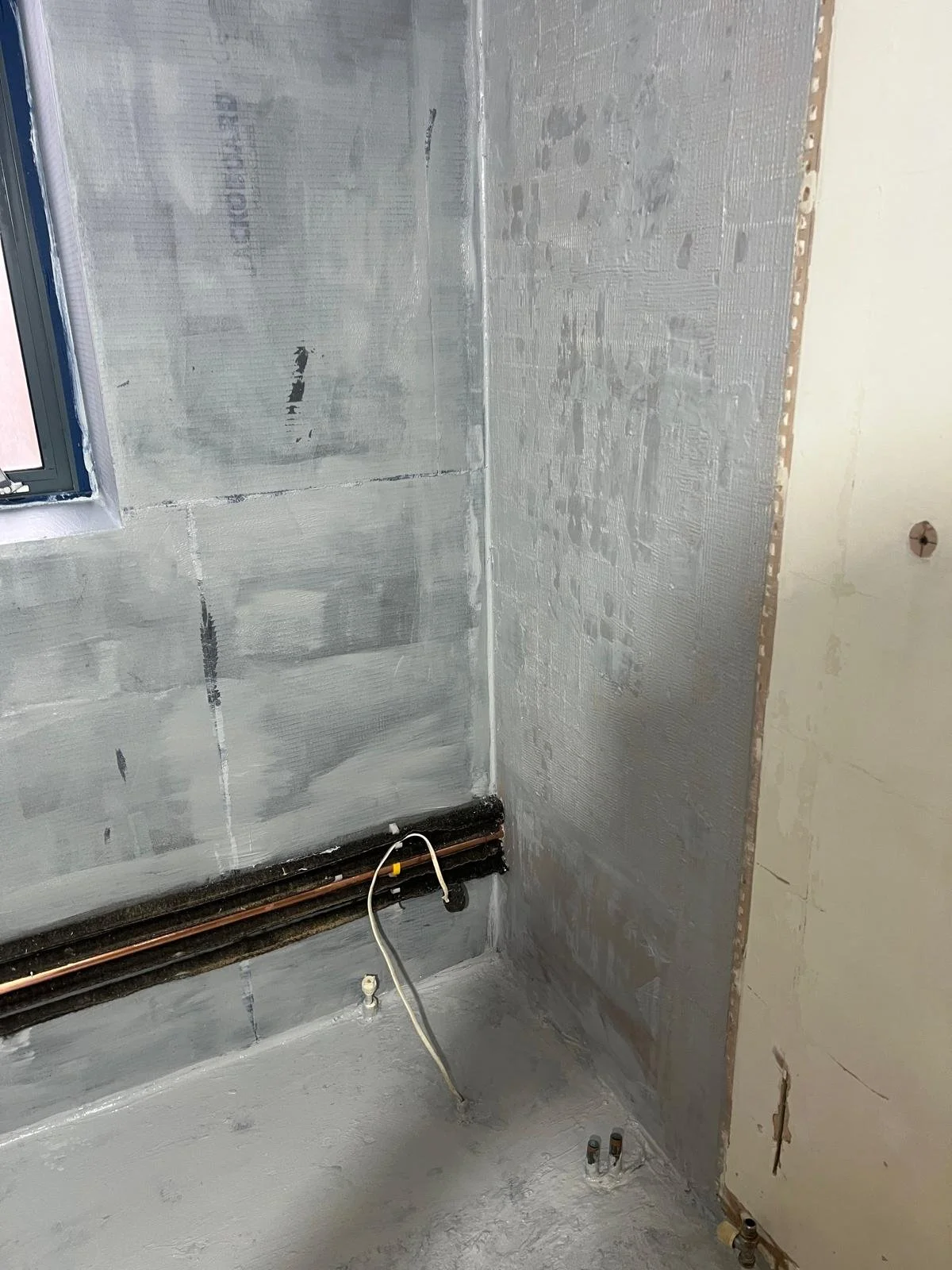 Interior view of a room under renovation with partially covered walls, exposed piping, and a window.