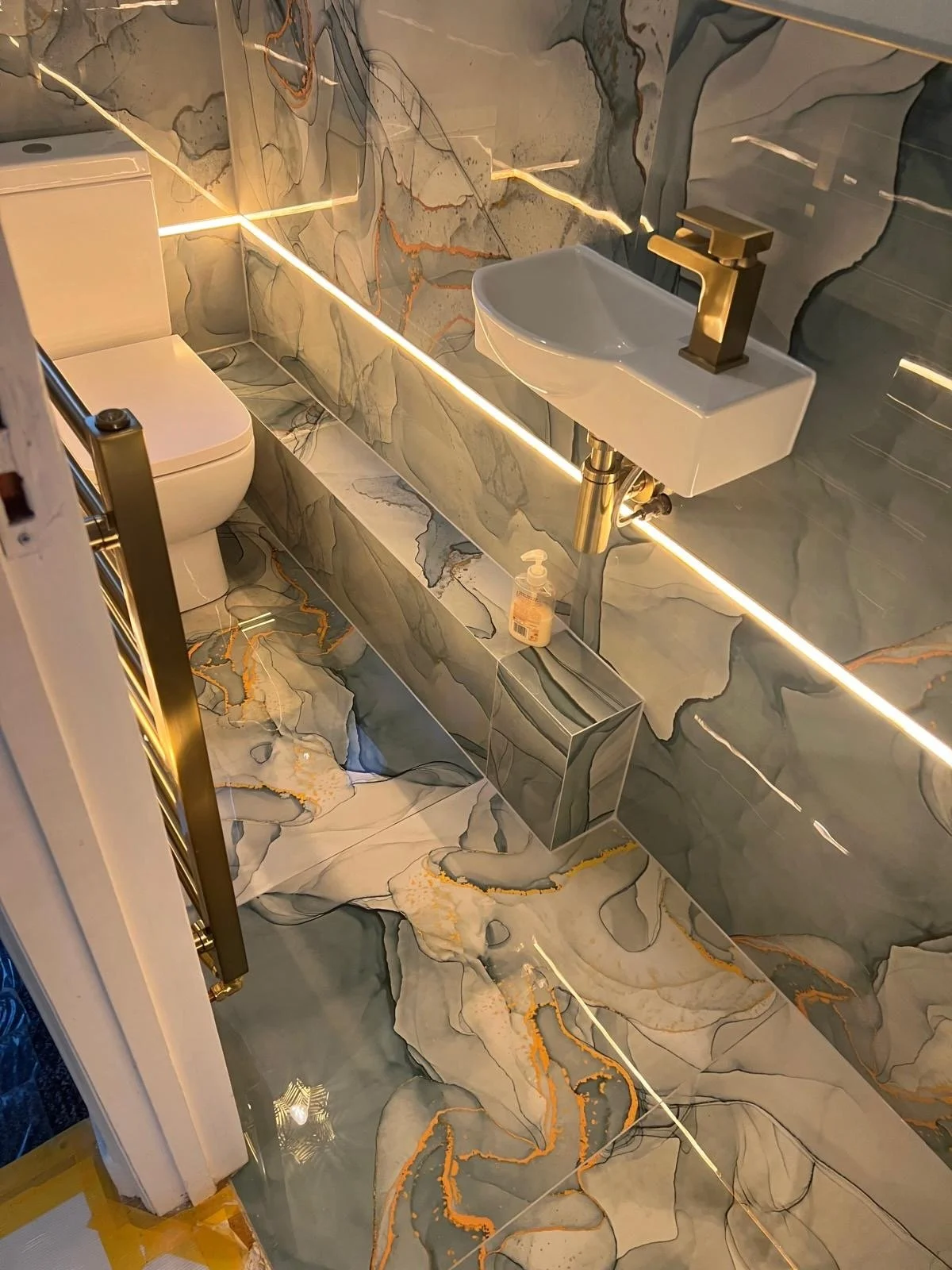 Bathroom with marble walls and floor, a toilet, a small white sink with a gold faucet, and LED strip lighting behind the sink and along the wall.