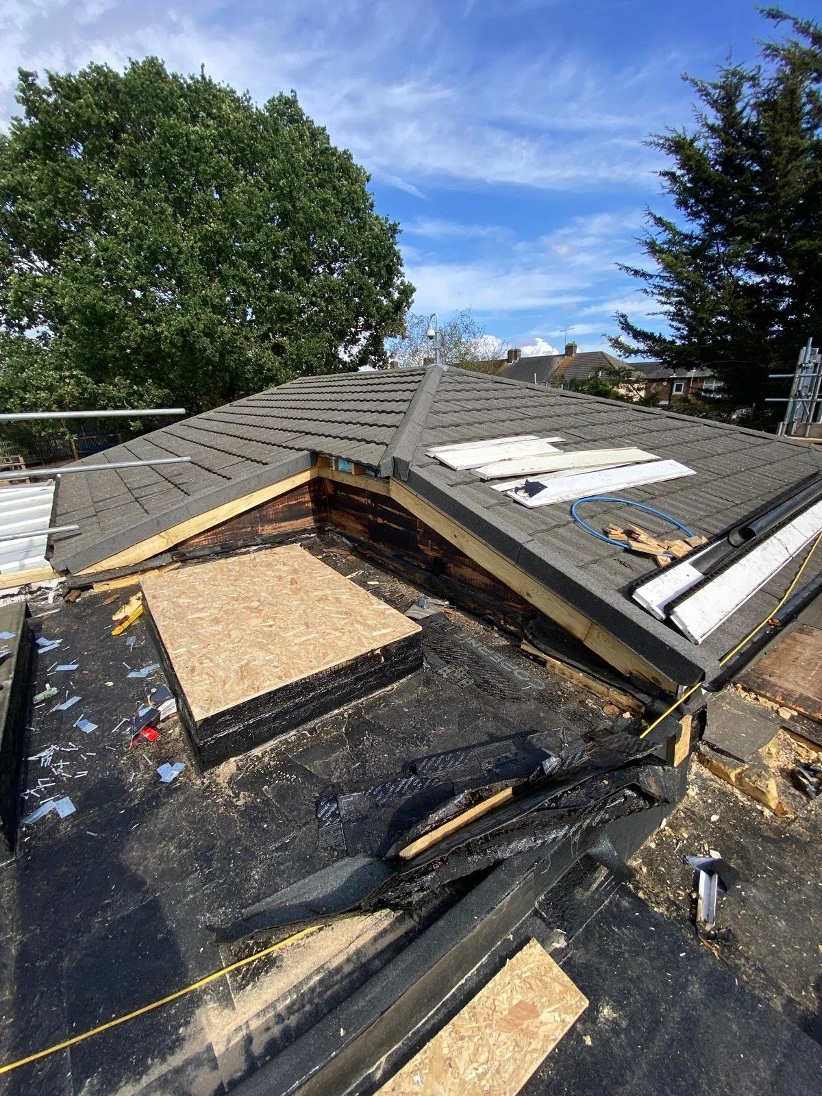 Roof under construction with tiles, plywood, and construction tools, some tile work is unfinished, trees and houses visible in the background.
