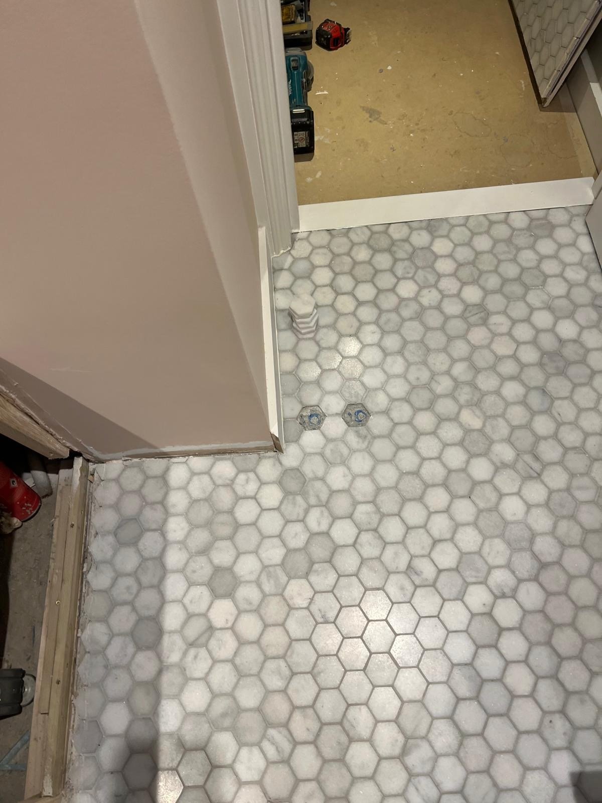 A kitchen floor with white hexagonal tiles and two visible plumbing bolts. The room is under renovation, with tools and construction materials visible in the adjacent room.