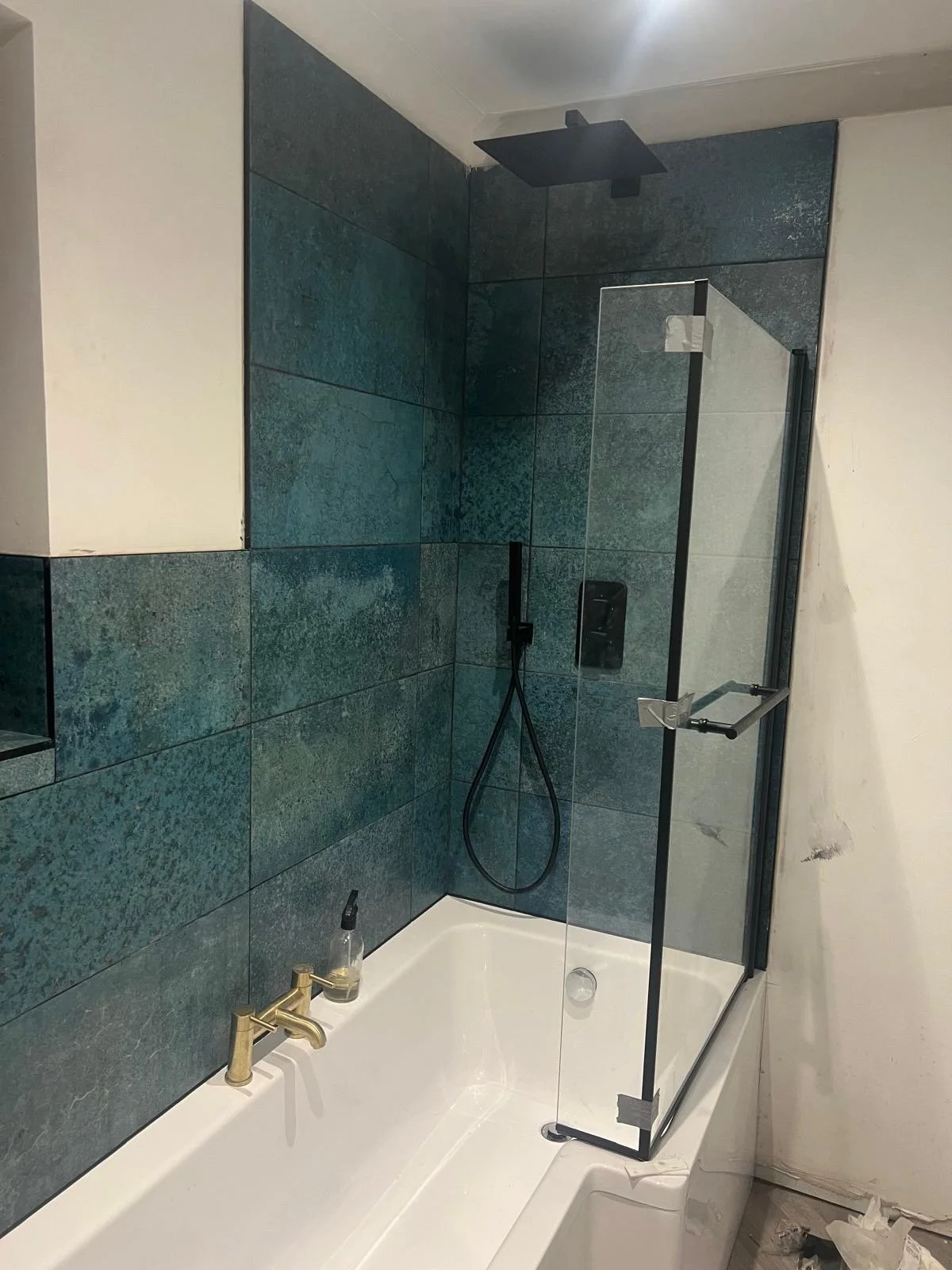 Modern shower area with dark green tiles, a black showerhead, a glass door, and a bathtub with a gold faucet.