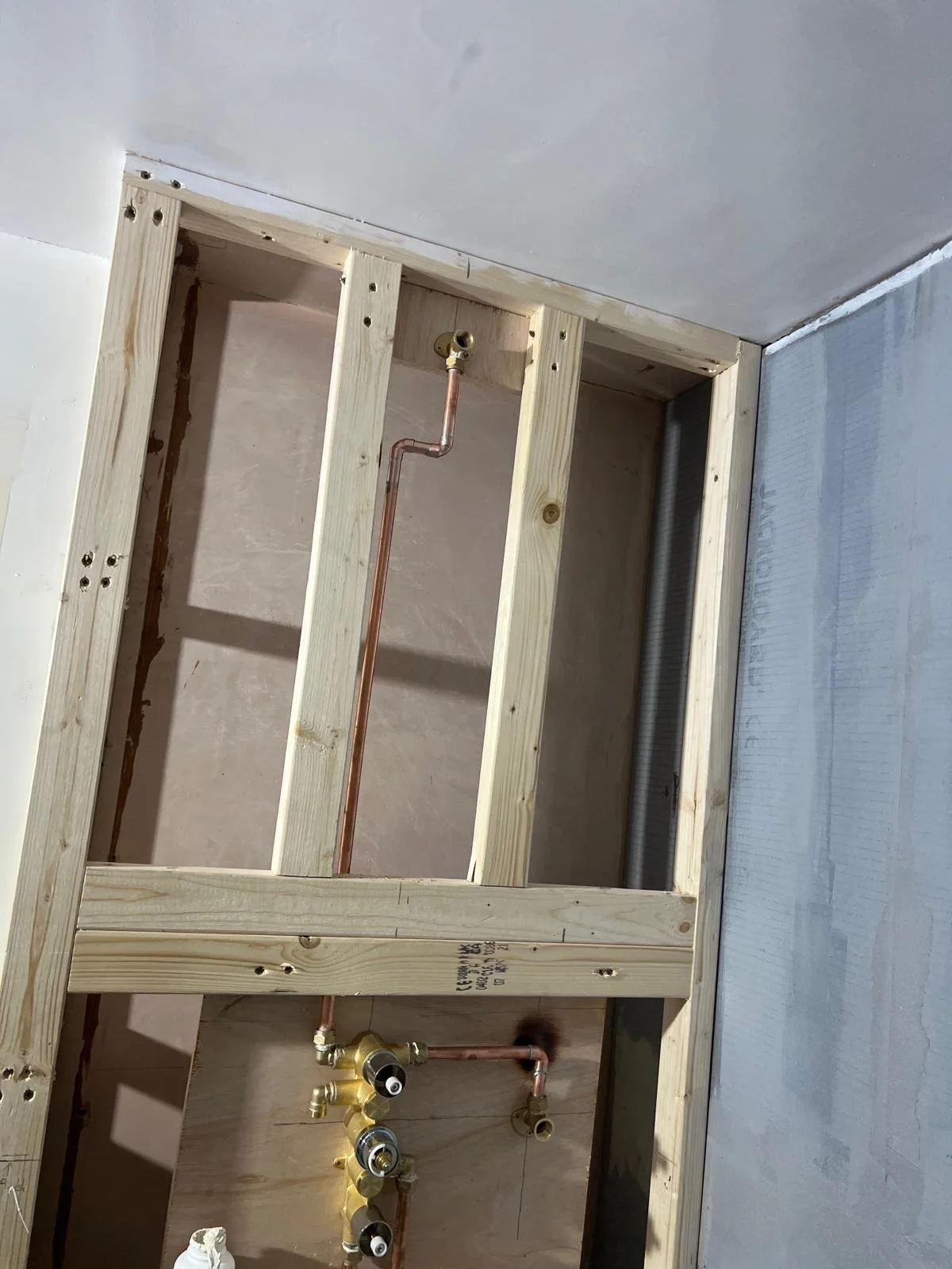 Interior view of a bathroom under construction showing wooden framing, copper pipes, and plumbing fixtures.