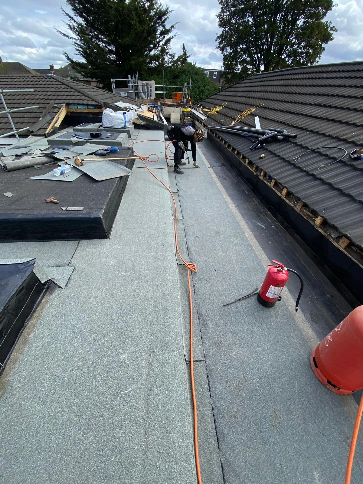 Roof under construction with worker installing or repairing roofing materials, safety equipment, and tools visible, including fire extinguisher.