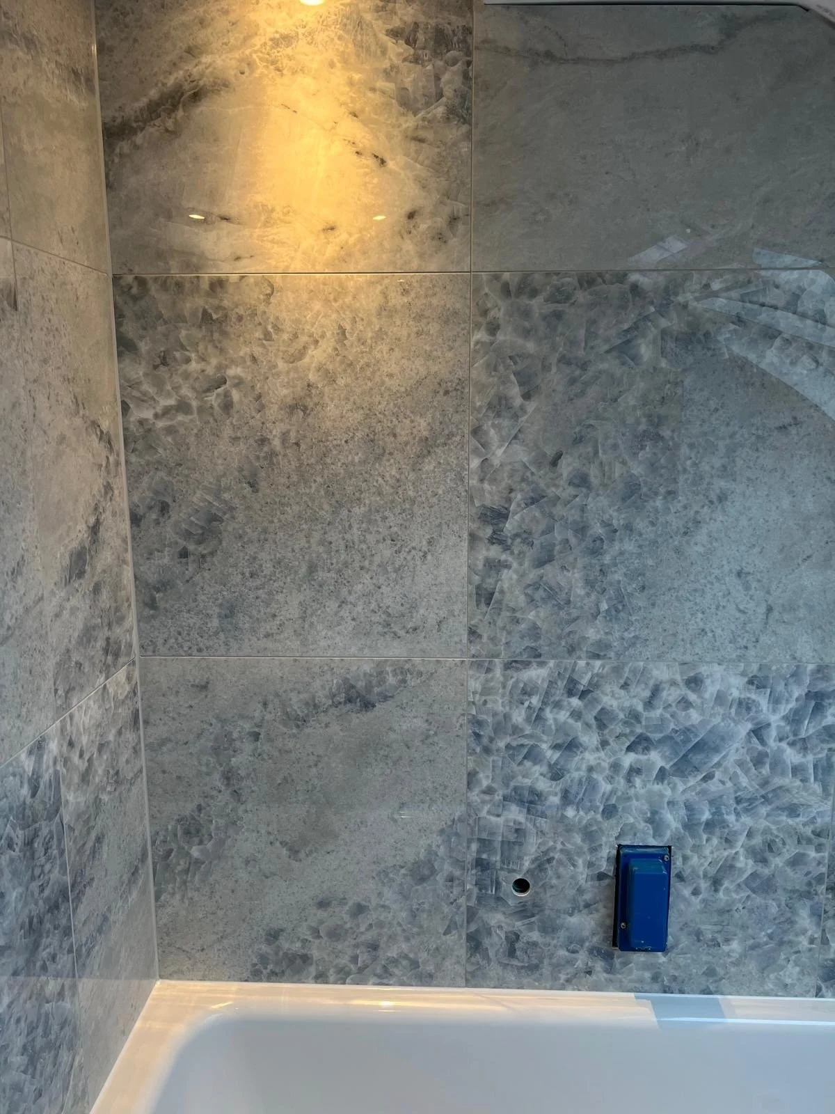 Interior view of a shower with grey marble tiles on the walls, a small round drain hole, and a blue box or bathroom fixture attached to the wall.