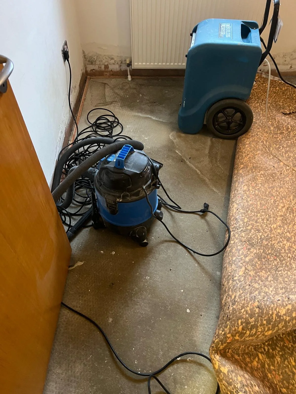 A vacuum cleaner and a carpet cleaning machine in a small room with a worn floor, a wall with peeling paint, and a radiator.