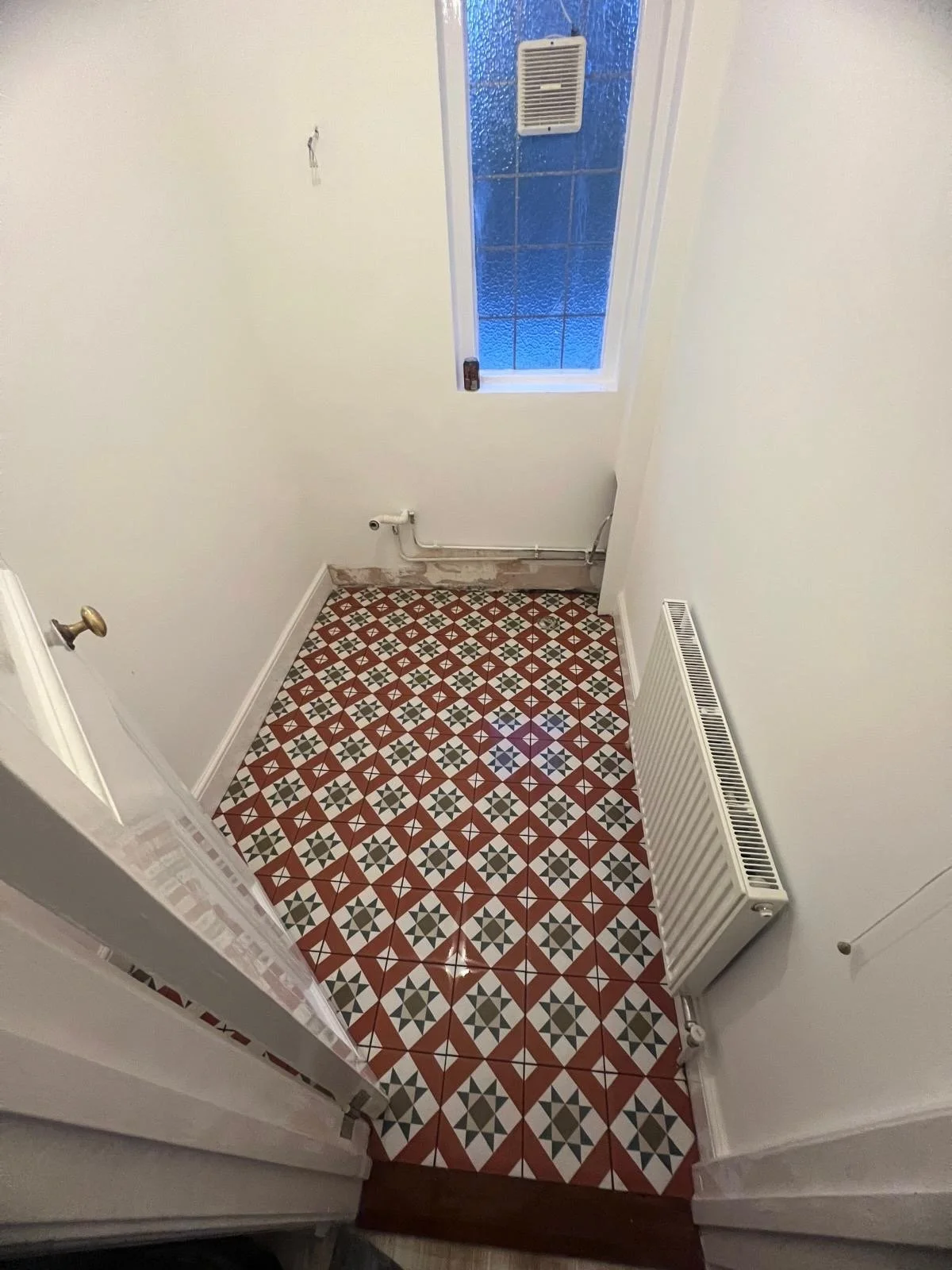 Empty small room or closet with mosaic tile floor, a white door with a gold knob, a radiator on the right wall, and a pipe running along the back wall, with a small window at the back covered in textured glass.