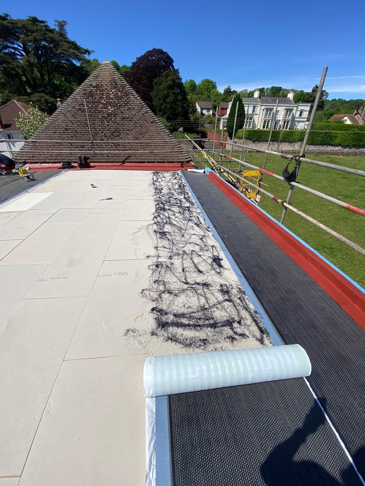 Roof under construction with white tiles, black dirt scattering, and scaffolding around the edge. In the background, neighboring houses and trees are visible under a clear blue sky.