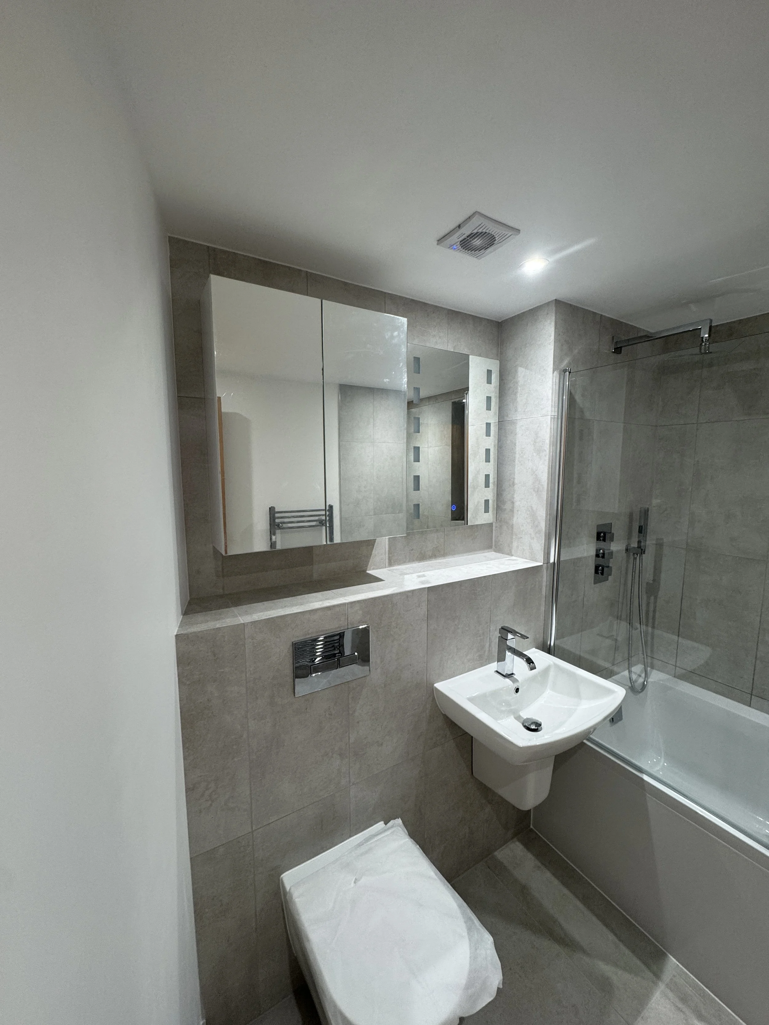 A modern bathroom featuring a wall-mounted mirror, a small rectangular sink with a chrome faucet, a wall-mounted toilet, a bathtub with a glass shower enclosure, and gray tiled walls. There is a towel heater mounted on the wall and an exhaust vent in