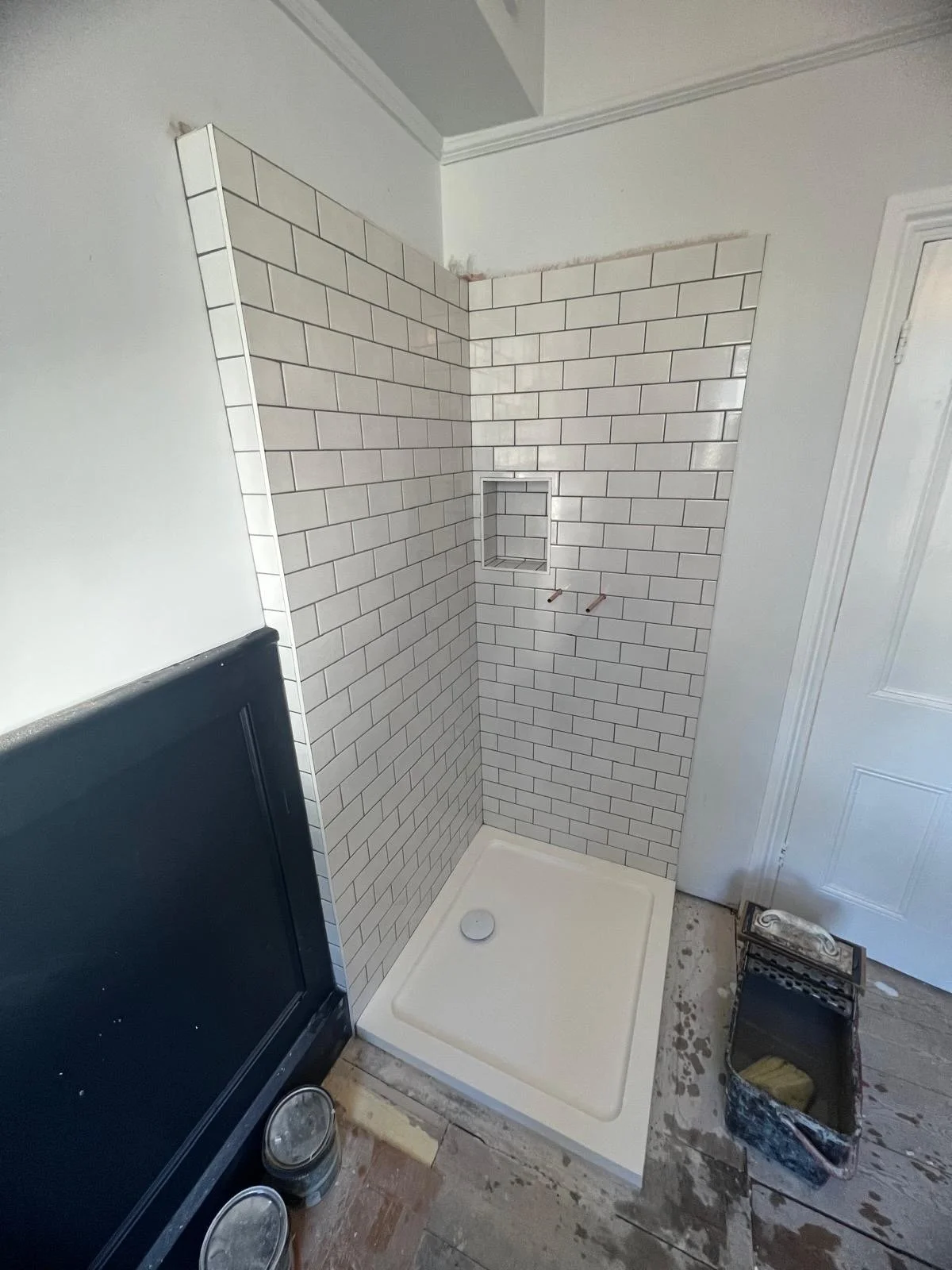 A bathroom corner under construction shows a partially tiled shower with white subway tiles, a built-in niche, and plumbing fixtures not yet installed. The shower base is in place, and construction tools and materials are nearby.