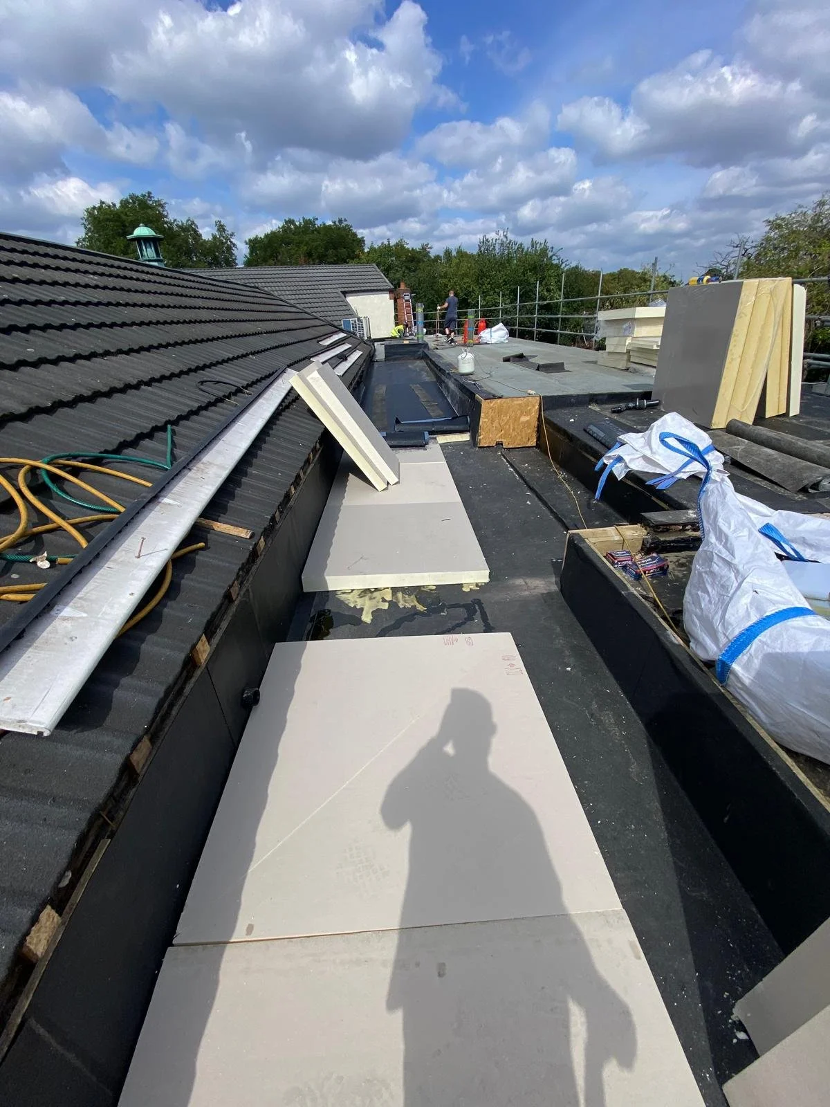 Construction workers installing a flat roof with tiles and insulation on a house, with scattered building materials and tools, under a partly cloudy sky.
