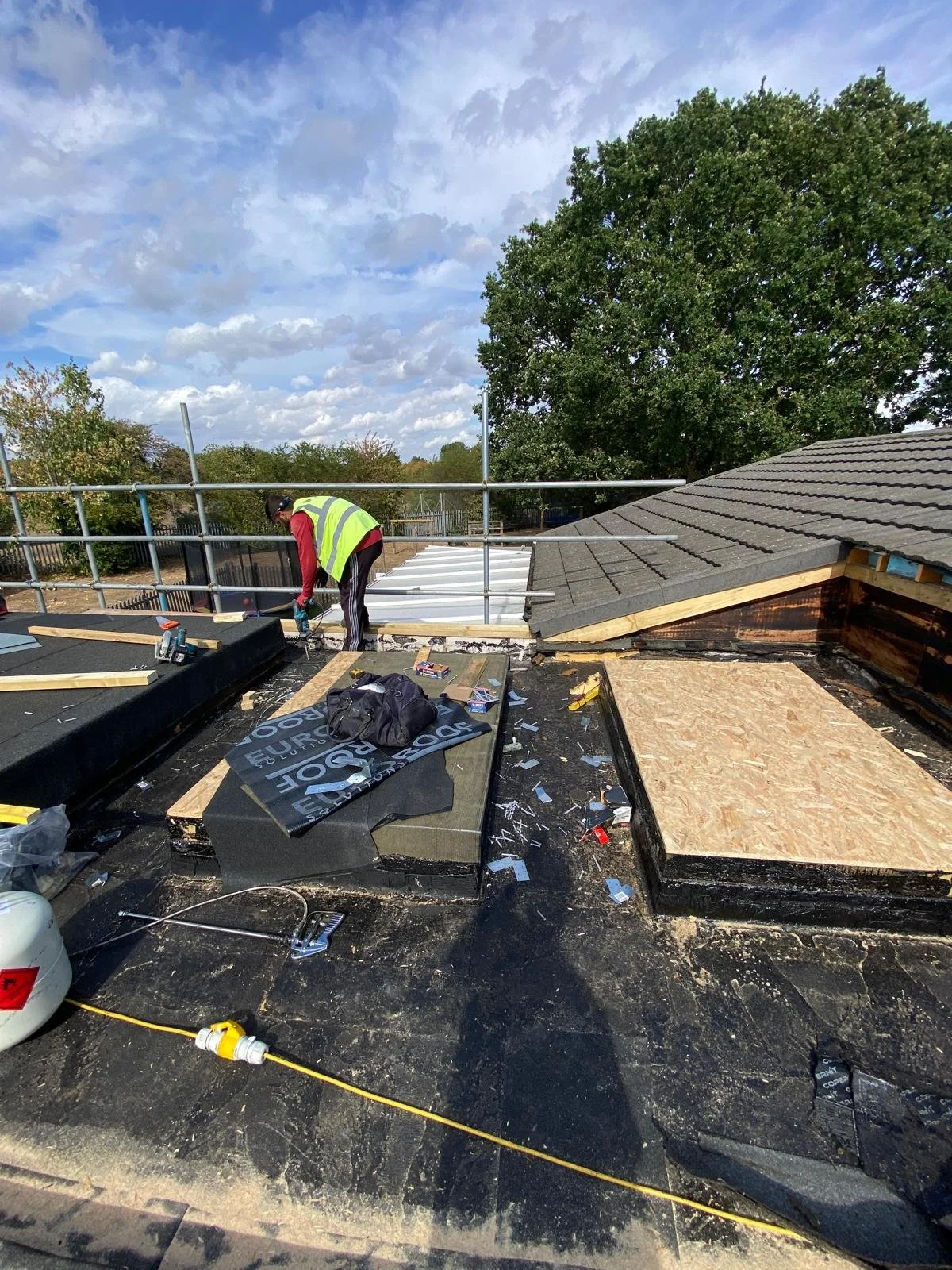 A construction worker in a high-visibility vest working on a roof under a cloudy sky, with trees and rooftops in the background.