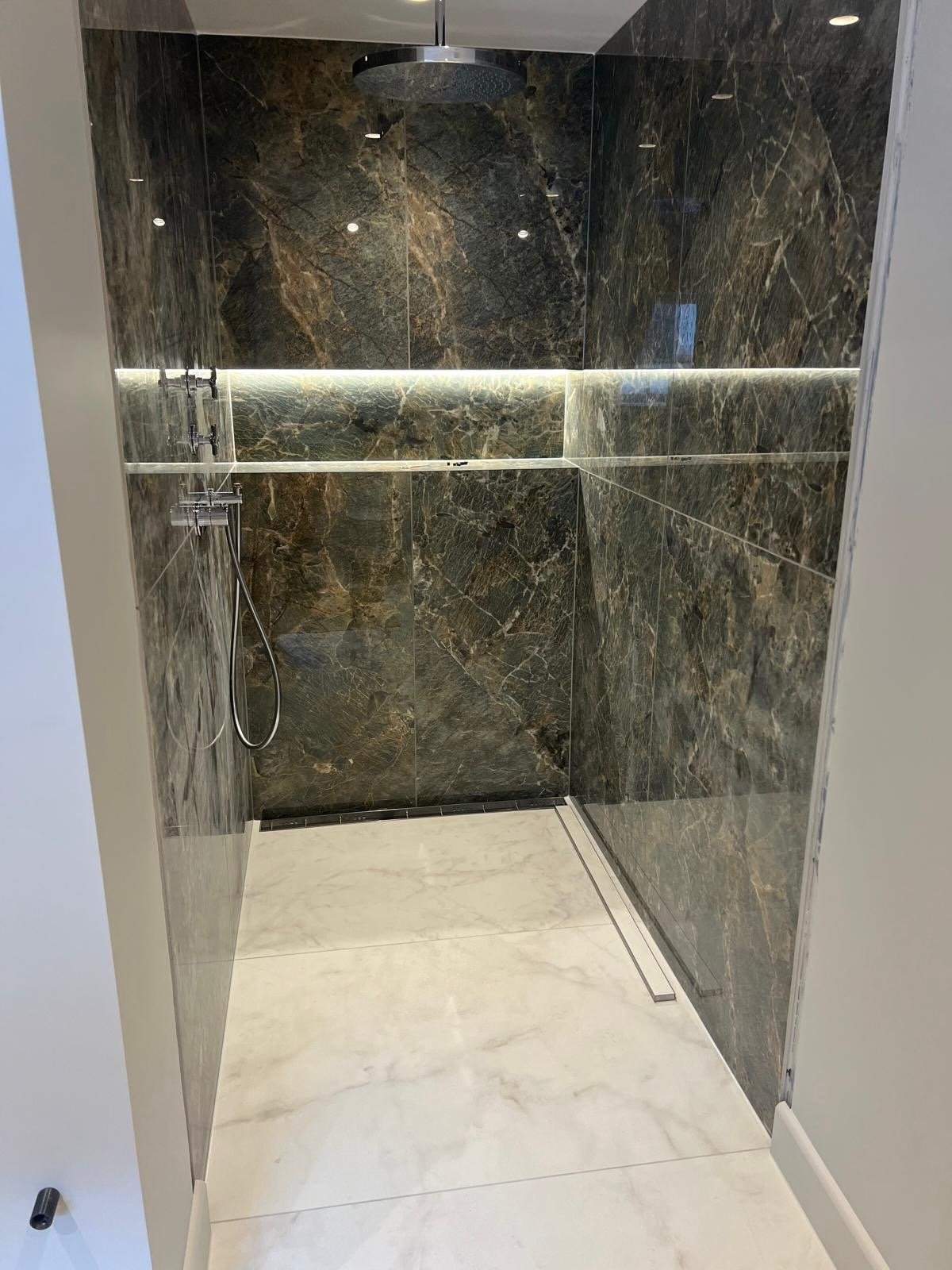 Modern walk-in shower with dark marble walls, a ceiling-mounted rain shower head, and a handheld shower on the left wall, illuminated by a horizontal LED light strip.