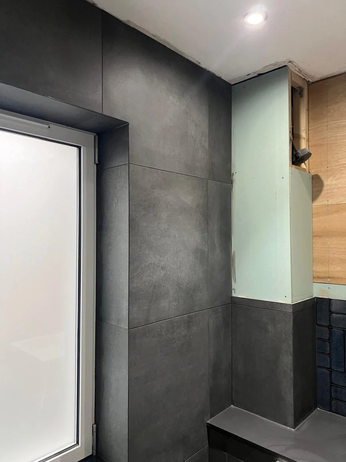 Interior view of a room under construction with a grey tiled wall, a partially installed green cabinet, and a wooden wall cabinet on the right. A recessed ceiling light is visible.