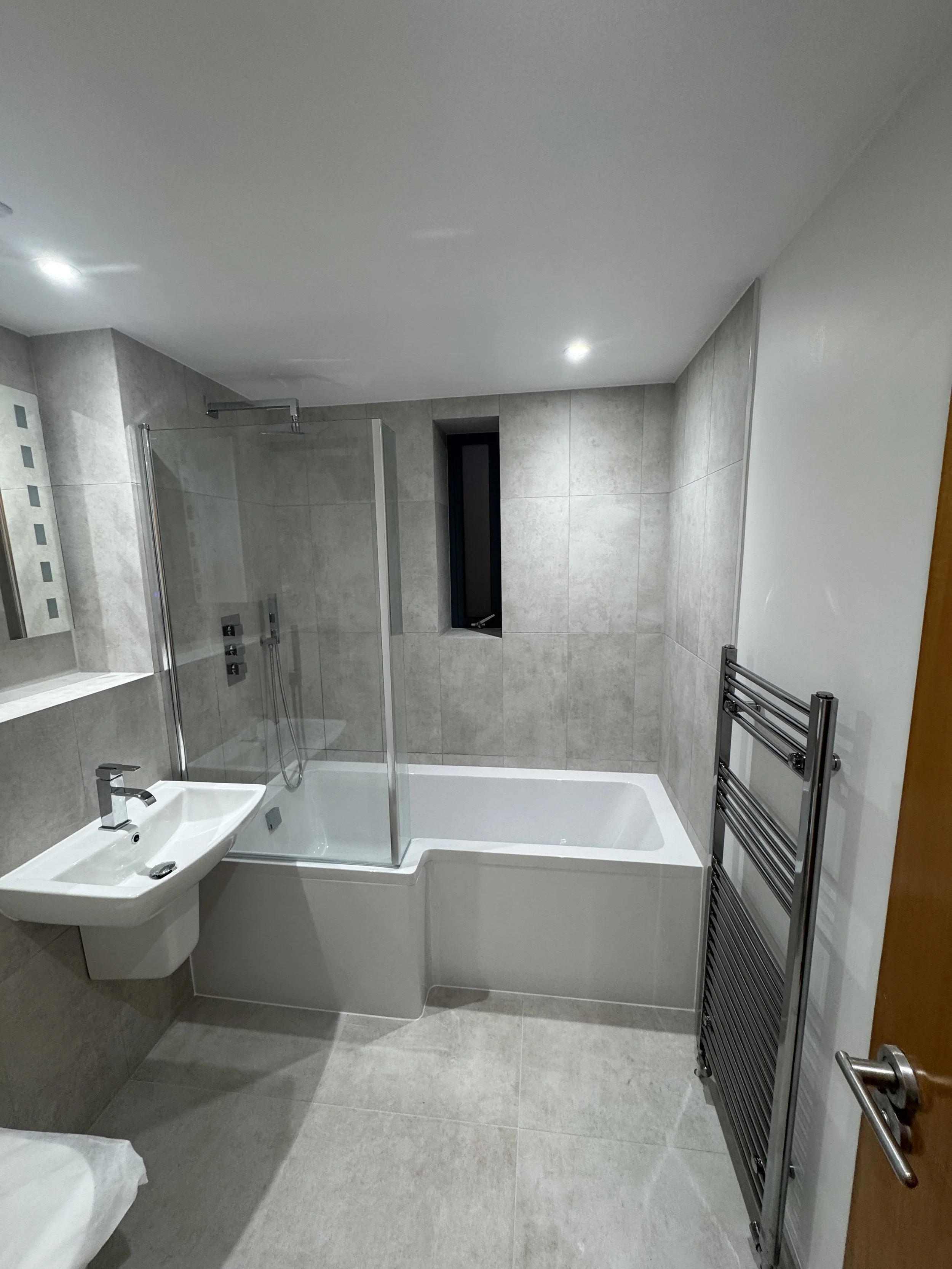 Modern bathroom with gray tiled walls and floor, featuring a bathtub with a glass shower screen, a small white sink, a window, and a wall-mounted heated towel rail.