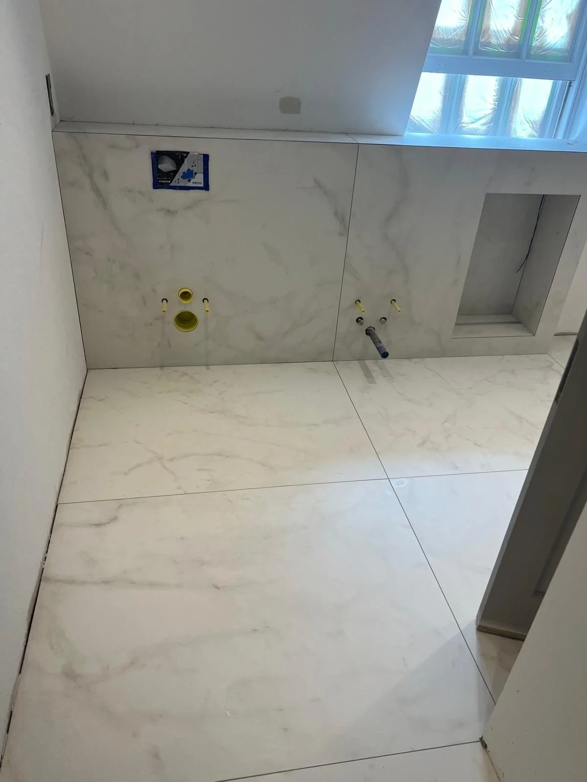 Bathroom under construction with marble walls and floor, plumbing pipes installed, and an opening for a mirror.