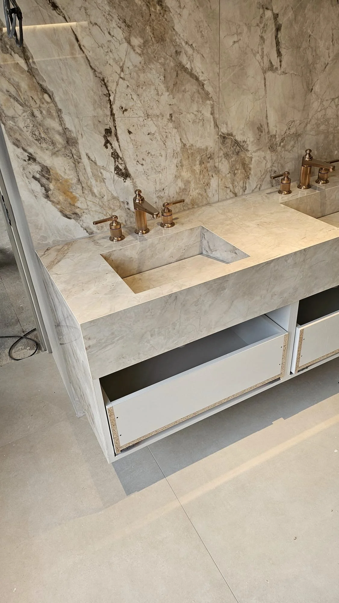 A marble double sink bathroom vanity with two bronze faucets and a marble backsplash. The vanity has open cabinet spaces below and is not fully assembled.