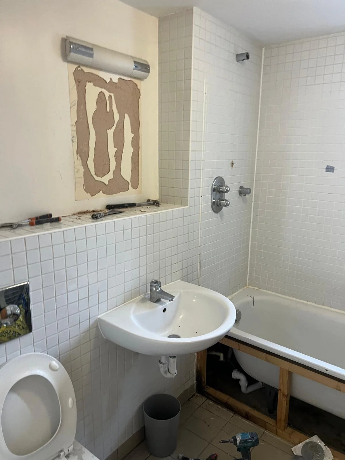 A bathroom under renovation with white tiled walls, a mounted light fixture, a small sink, an unfinished bathtub, and various tools and materials scattered around.