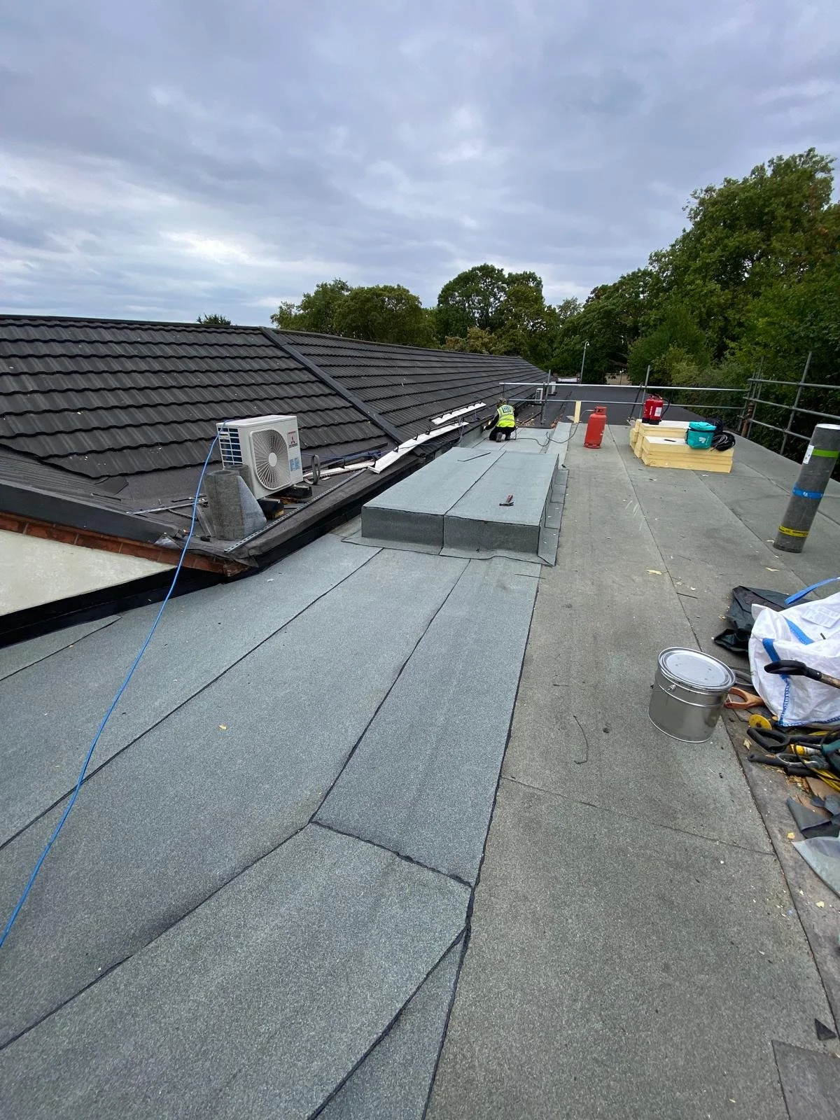 On a flat rooftop, two workers are installing or repairing roofing materials. There are construction tools and supplies scattered around, including a silver bucket, a red fire extinguisher, and stacked insulation. The sky is cloudy with some tree top