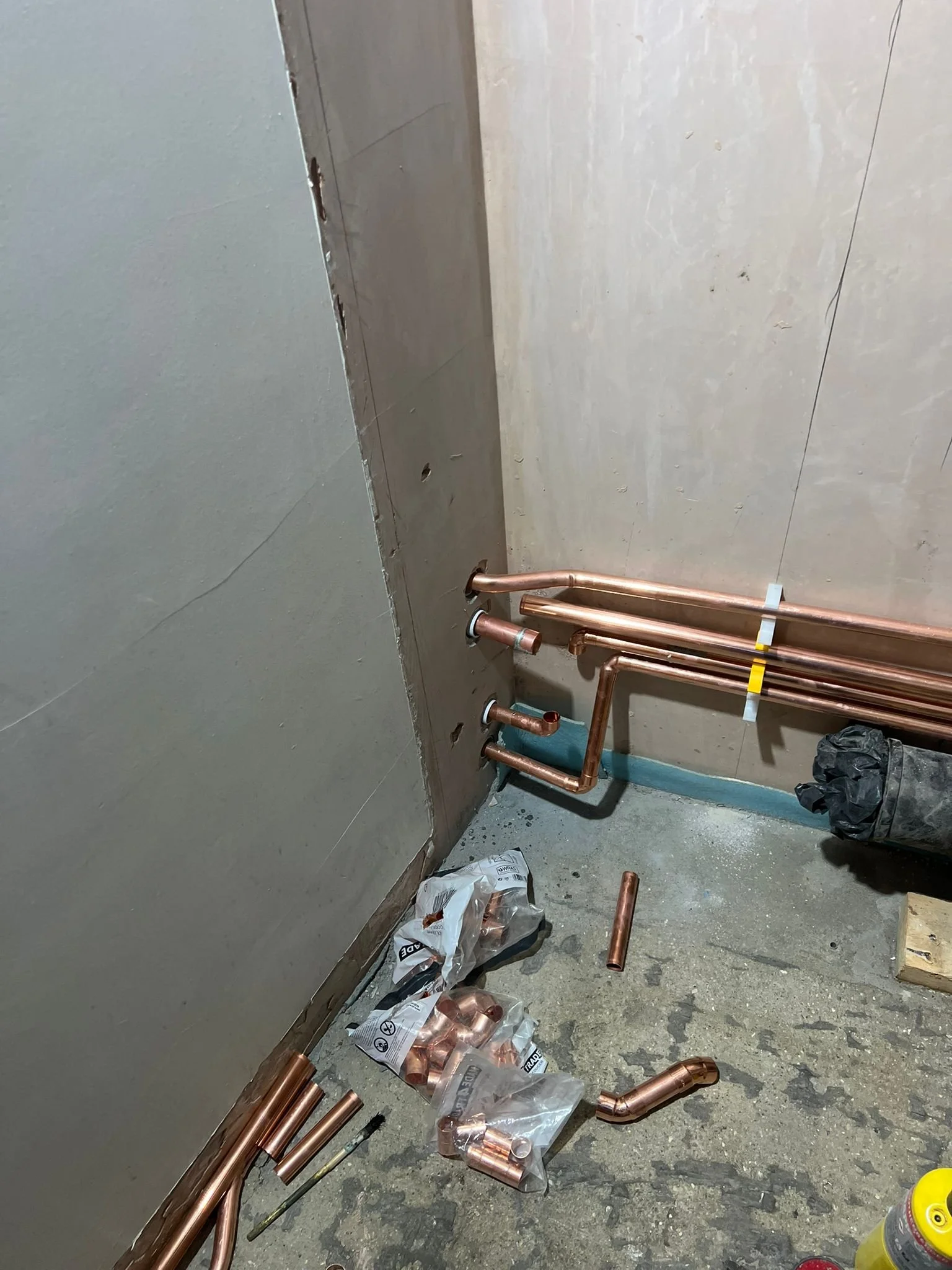 A construction site with copper pipes and fittings on the floor and partially installed along the wall. There are plastic bags and tools scattered around, with a gray wall and concrete floor.