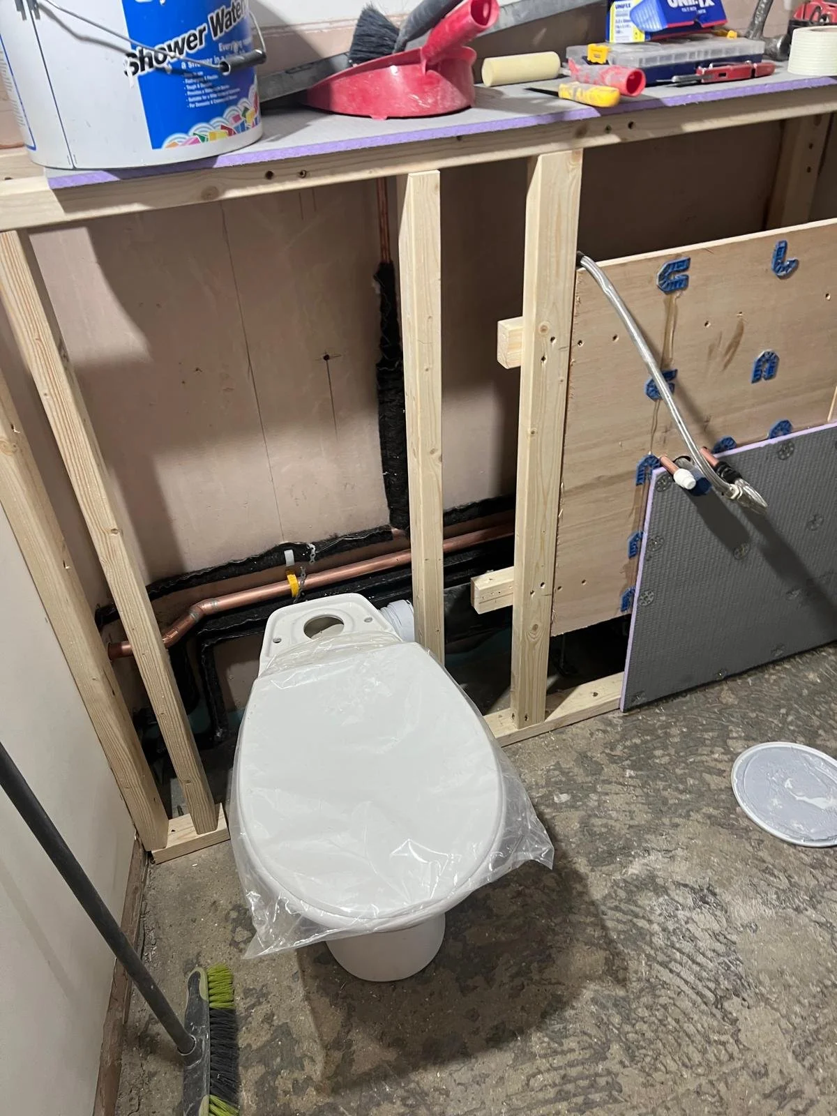 A partially constructed bathroom with exposed wooden framing, plumbing, and a toilet wrapped in plastic. Tools and materials are on the work surface above.