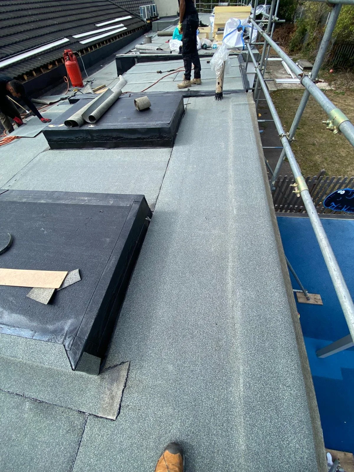 Construction workers on a roof installing a flat roofing system, with tools and materials scattered around, surrounded by safety scaffolding.