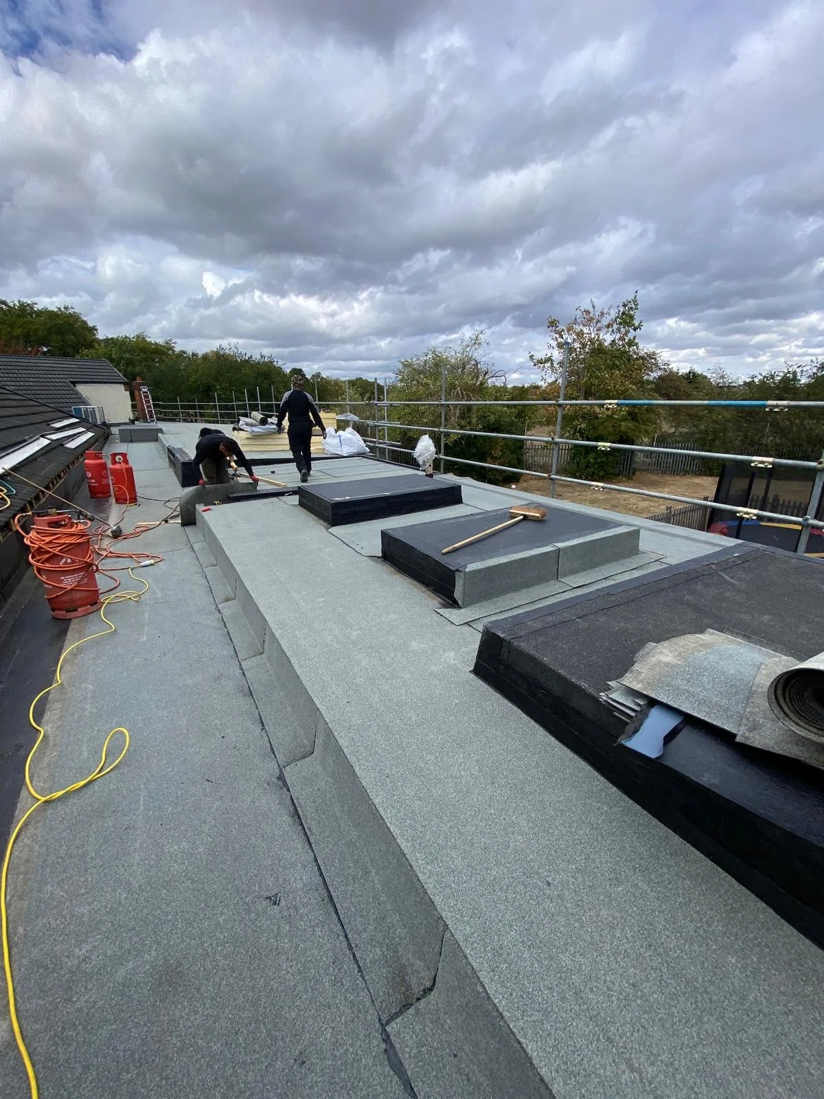 Two workers on a roof installing or repairing roofing materials under cloudy skies. Various tools and supplies are scattered around.