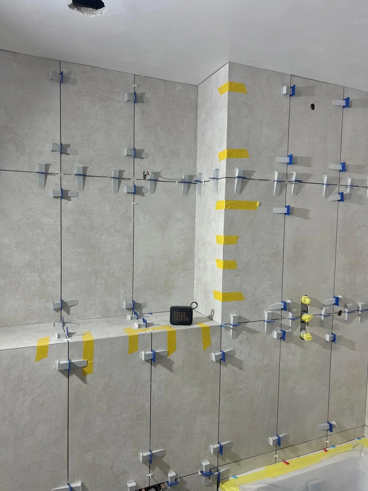Installing large beige tiles on a wall with blue and yellow spacers and tape, with a small JBL speaker placed on a ledge.