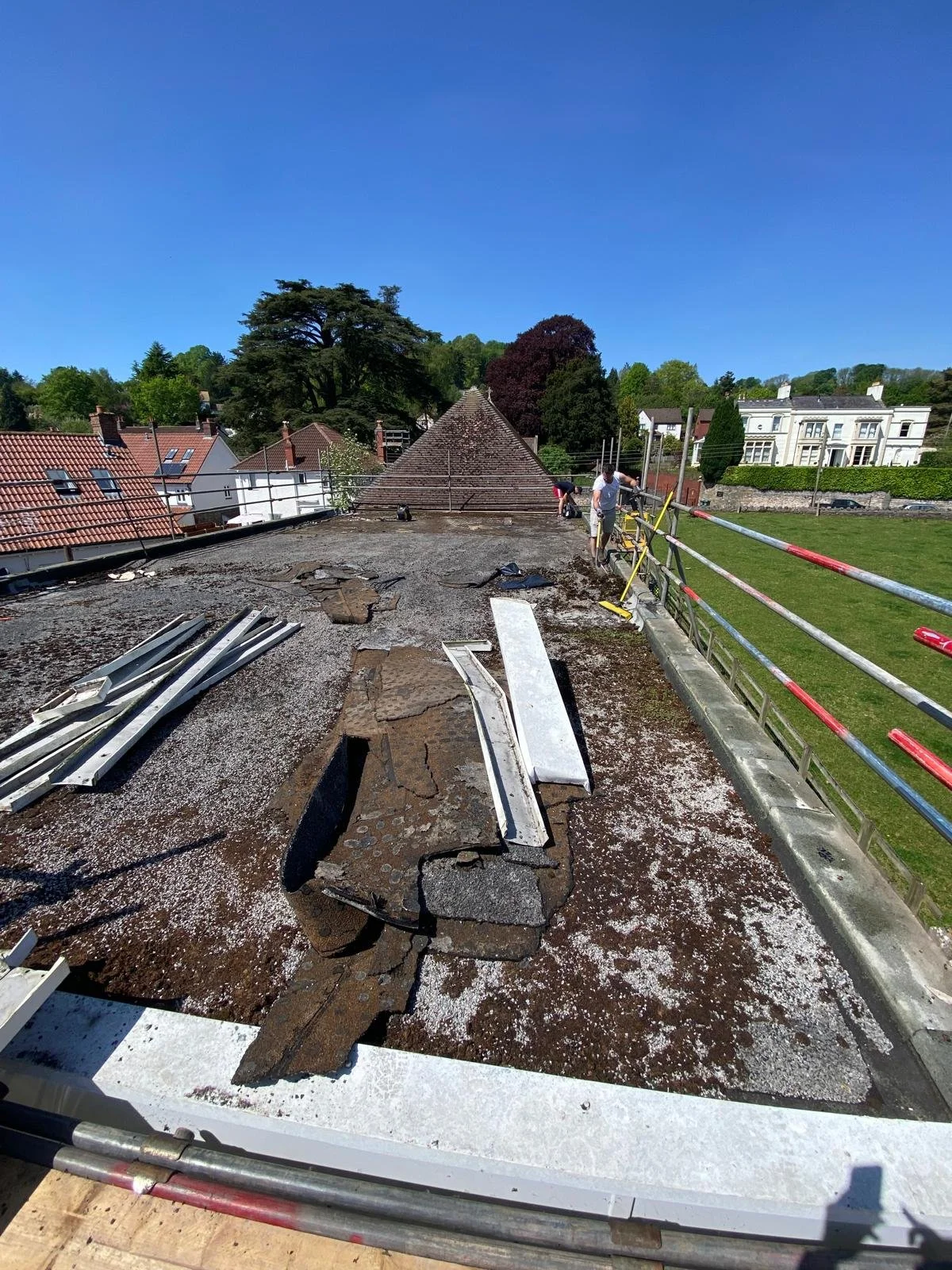 Construction workers are repairing the roof of a building, with roofing materials and debris scattered across the surface, surrounded by metal scaffolding and a green lawn.