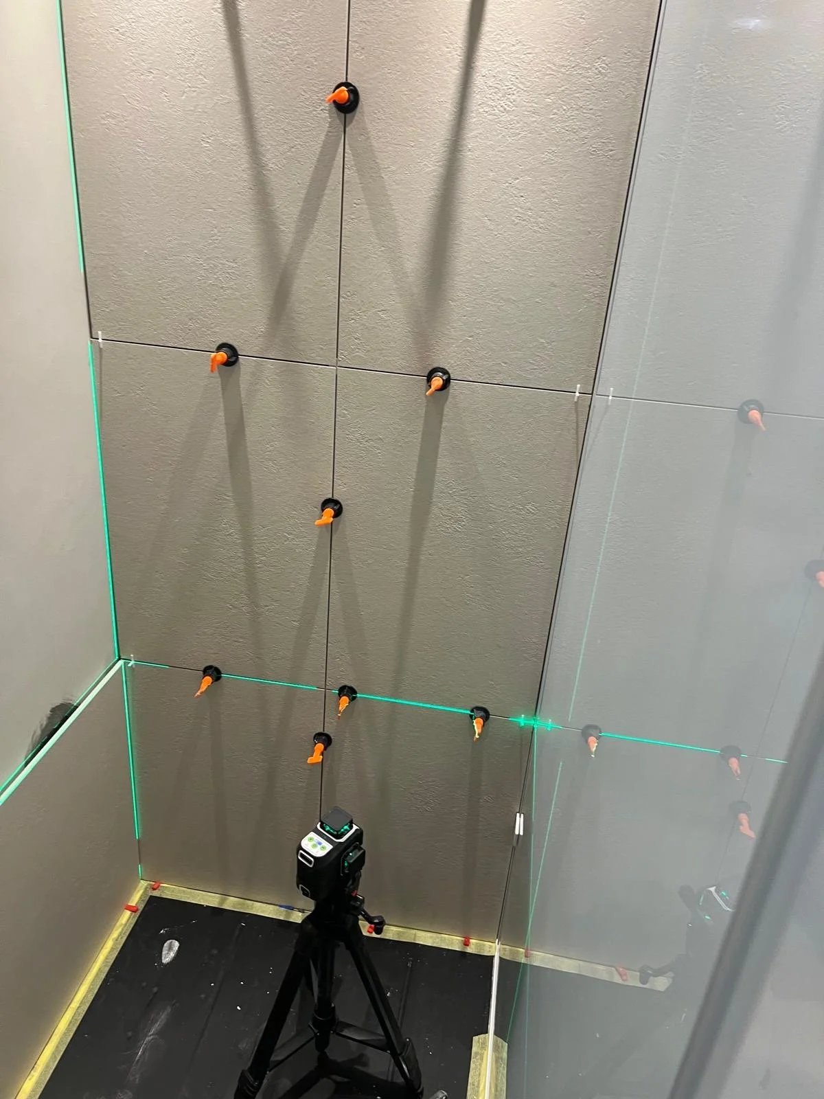 A room with gray walls and a gray ceiling, equipped with four orange and black laser triangle targets, a black tripod with a measuring device, and reflecting surfaces to test laser accuracy.