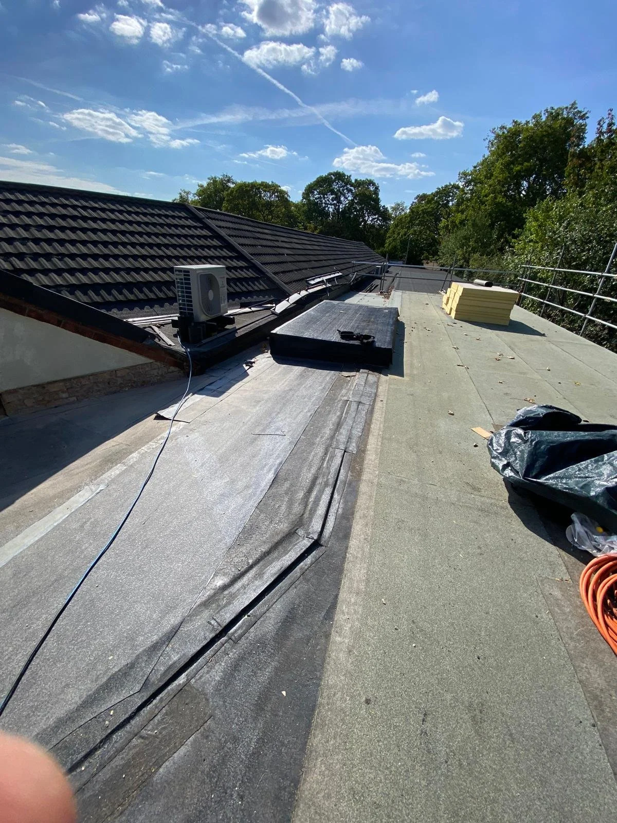 Construction site on a sloped roof with roofing work in progress, roofing materials, and an air conditioning unit under a partly cloudy sky.
