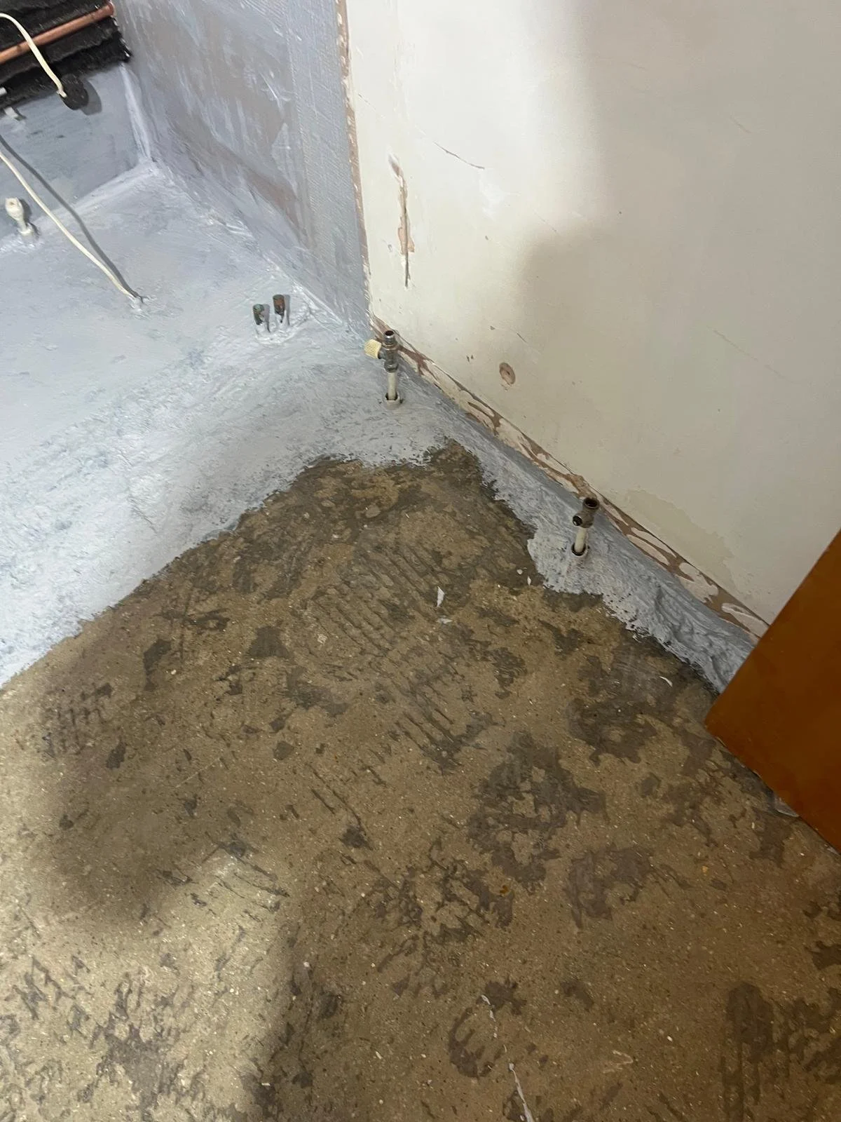 A room under renovation with a concrete subfloor and a wall with plumbing pipes protruding for a future fixture. The corner and base of the wall show exposed drywall and a partially finished coat of primer or paint.