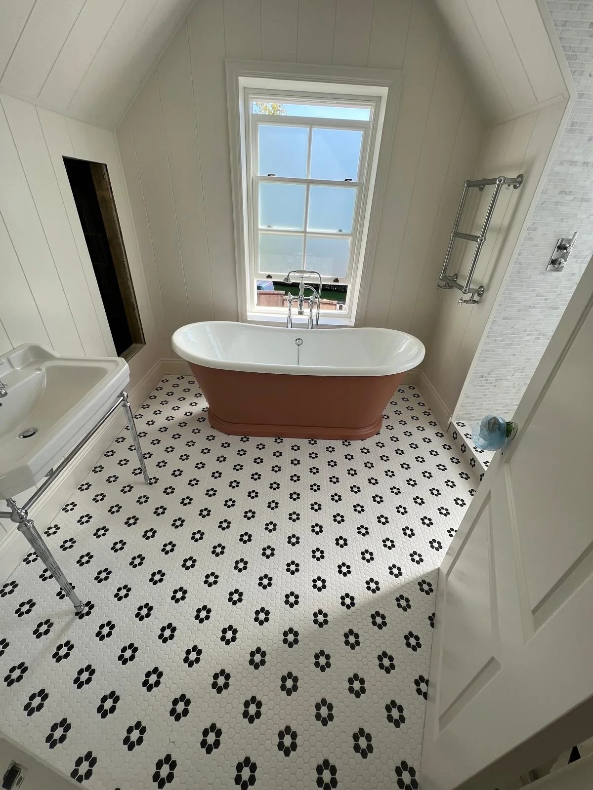 A bathroom with a vintage clawfoot bathtub placed in front of a large window, black and white floral patterned tile floor, white paneled walls, and a small sink to the left.