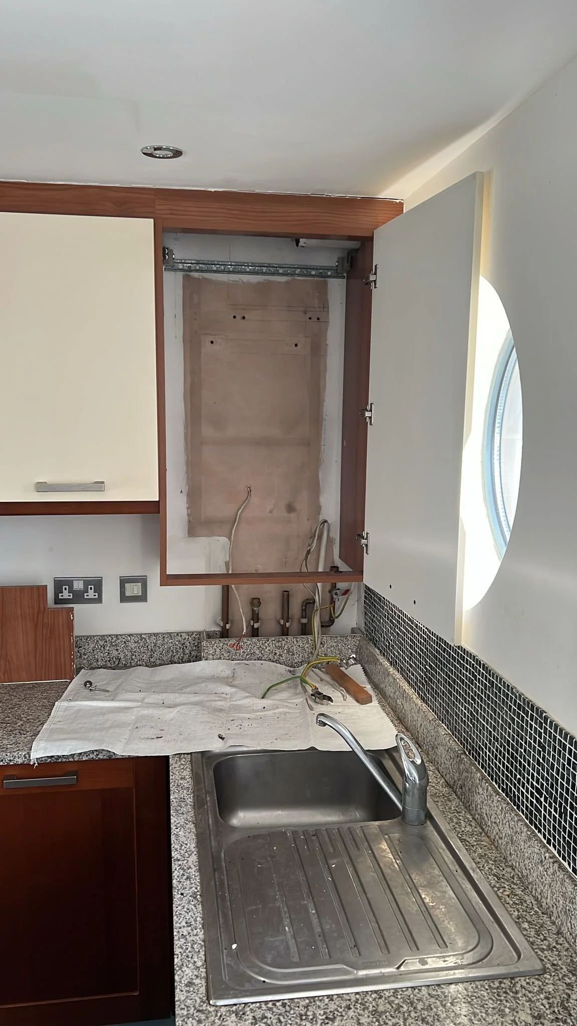Empty kitchen space with open cabinet above the sink, exposing plumbing and electrical wiring, sink with a stream of water running, granite countertop, and a window on the right side.