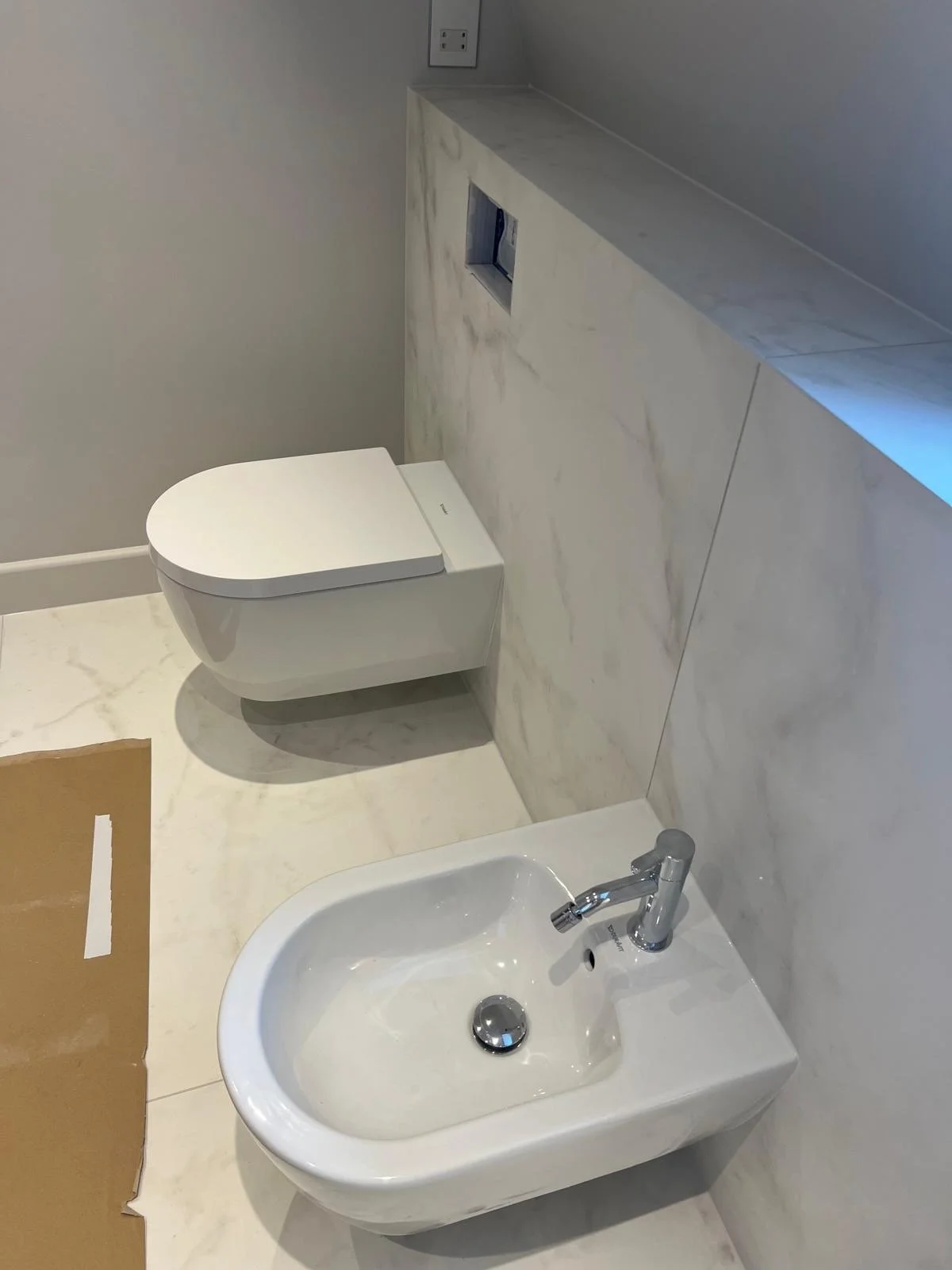 A bathroom with a white toilet and a bidet, both mounted on a marble-tiled wall and floor. A sink is also visible in the foreground.