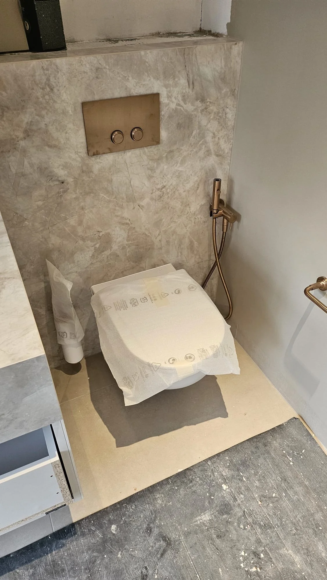 A toilet in a bathroom with a marble wall behind it, covered with protective paper. There is a small handheld bidet next to the toilet and a toilet paper roll on the floor.