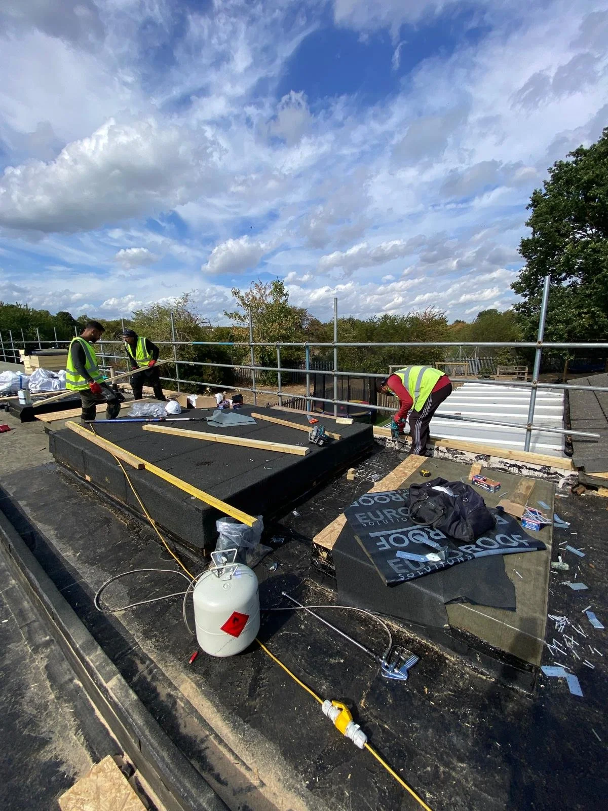 Construction workers wearing yellow safety vests and helmets working on a roof with tools and materials under a partly cloudy sky.