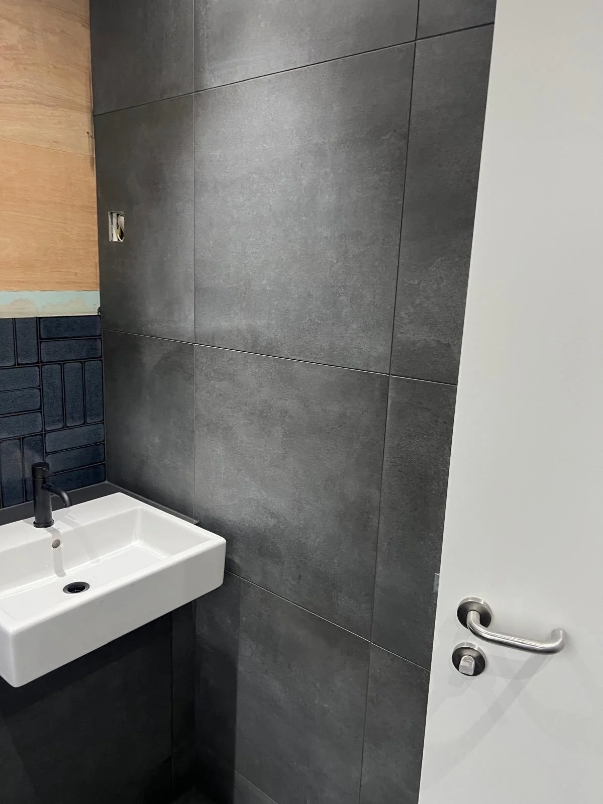 A small white sink with a black faucet mounted on a black cabinet, gray tiles on the wall behind it, with a section of blue tile and a wooden panel above, and a white door with a silver handle on the right side.