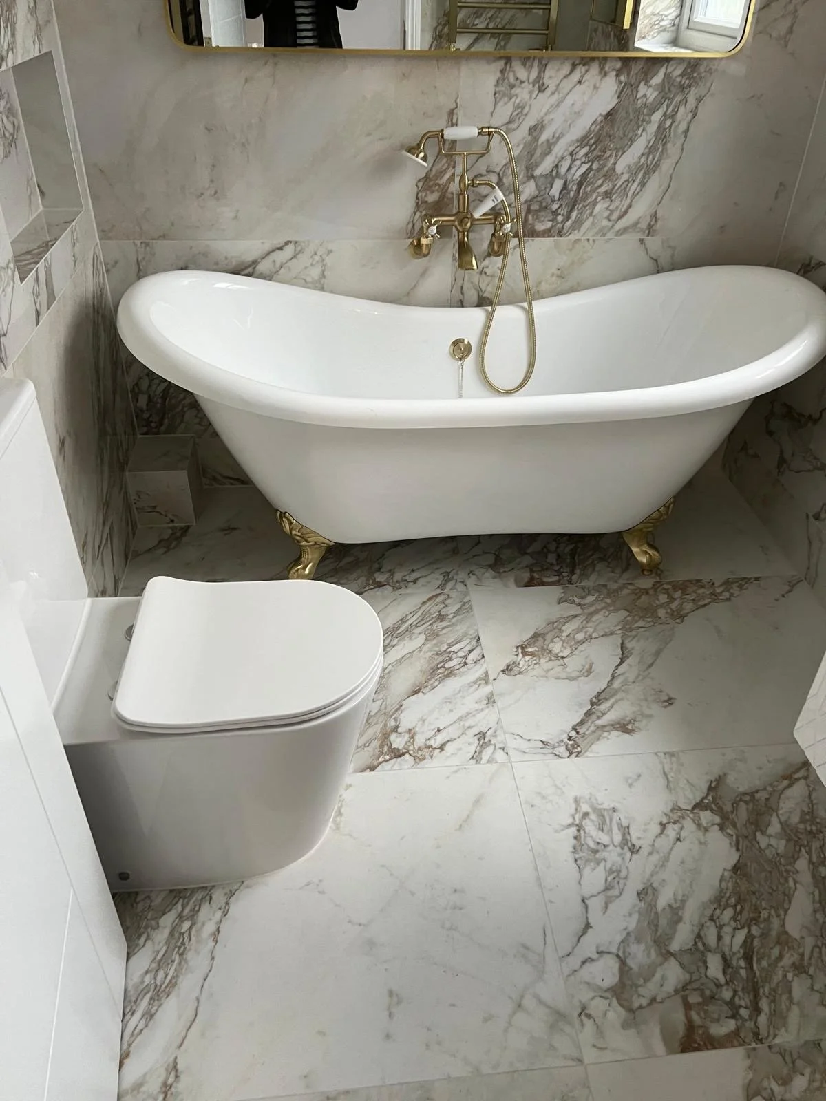 Bathroom with a classic clawfoot bathtub with gold accents, a white toilet, and marble tiles on the floor and walls.
