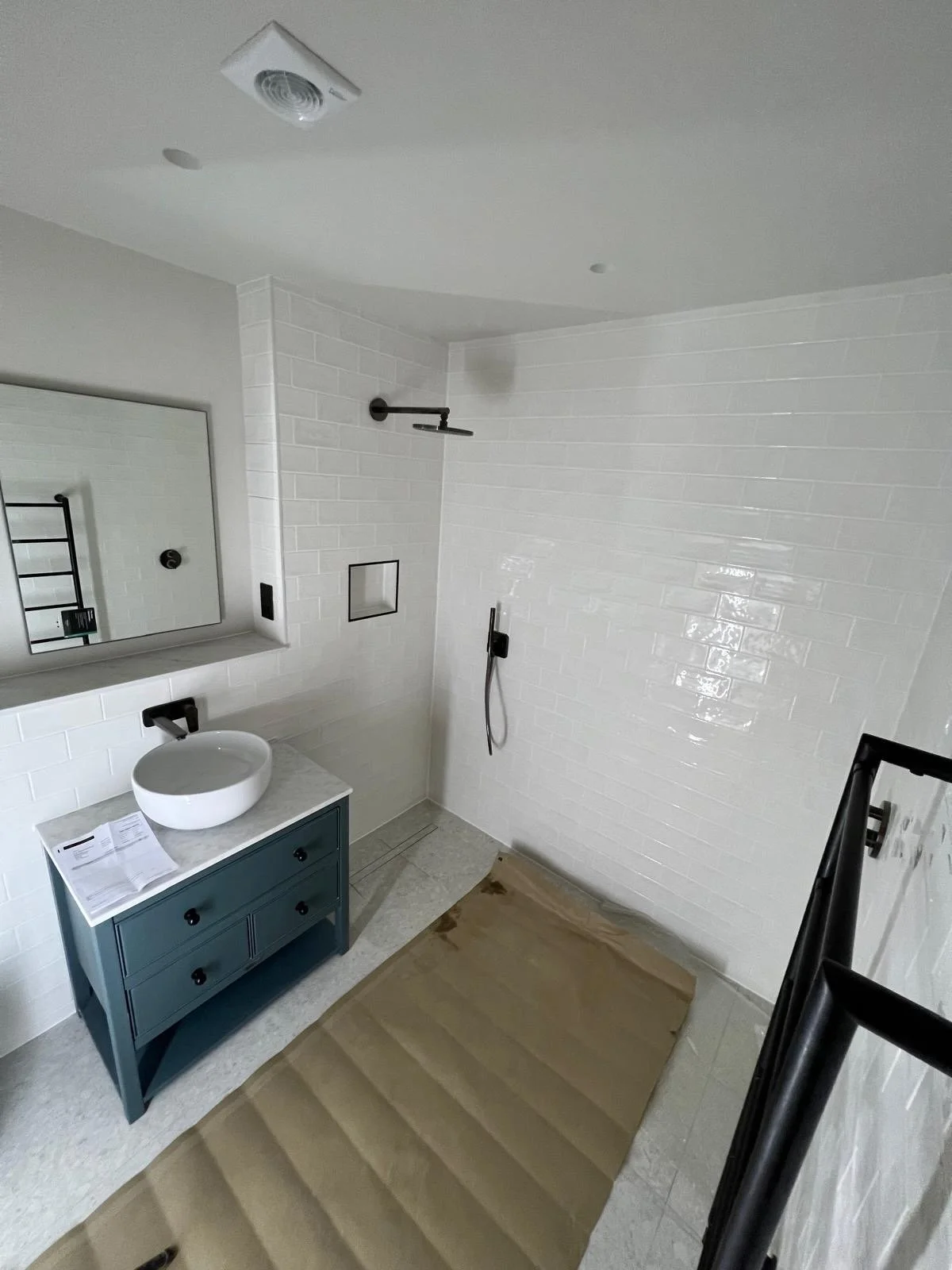 Bathroom under construction with white tiled shower, blue vanity with vessel sink, wall-mounted faucet, mirror, and black fixtures.