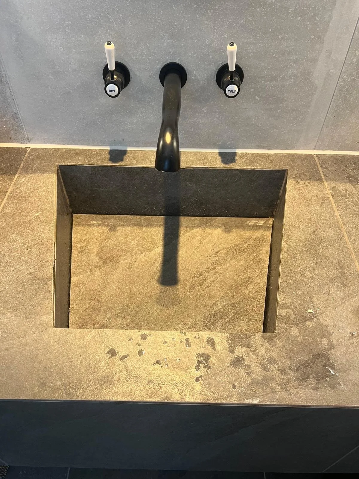 A modern bathroom sink with a black faucet and two handles labeled 'HOT' and 'COLD', above a square opening in the counter for a recessed basin.