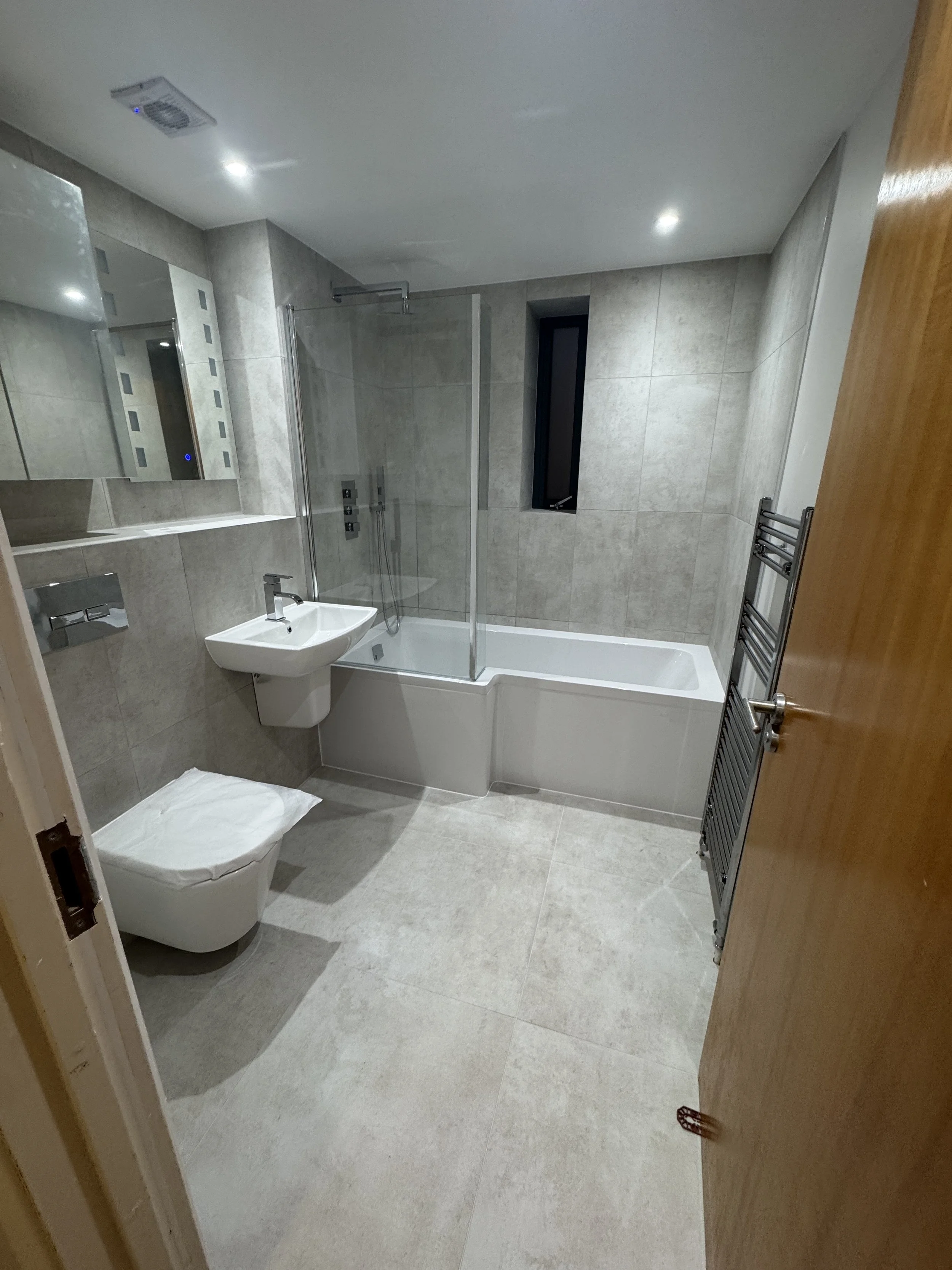 Modern bathroom with beige tiled walls and floor, a white toilet with a cloth on top, a small white sink, a mirrored cabinet, a glass shower enclosure, a narrow black window, metallic towel radiator, and wooden door.