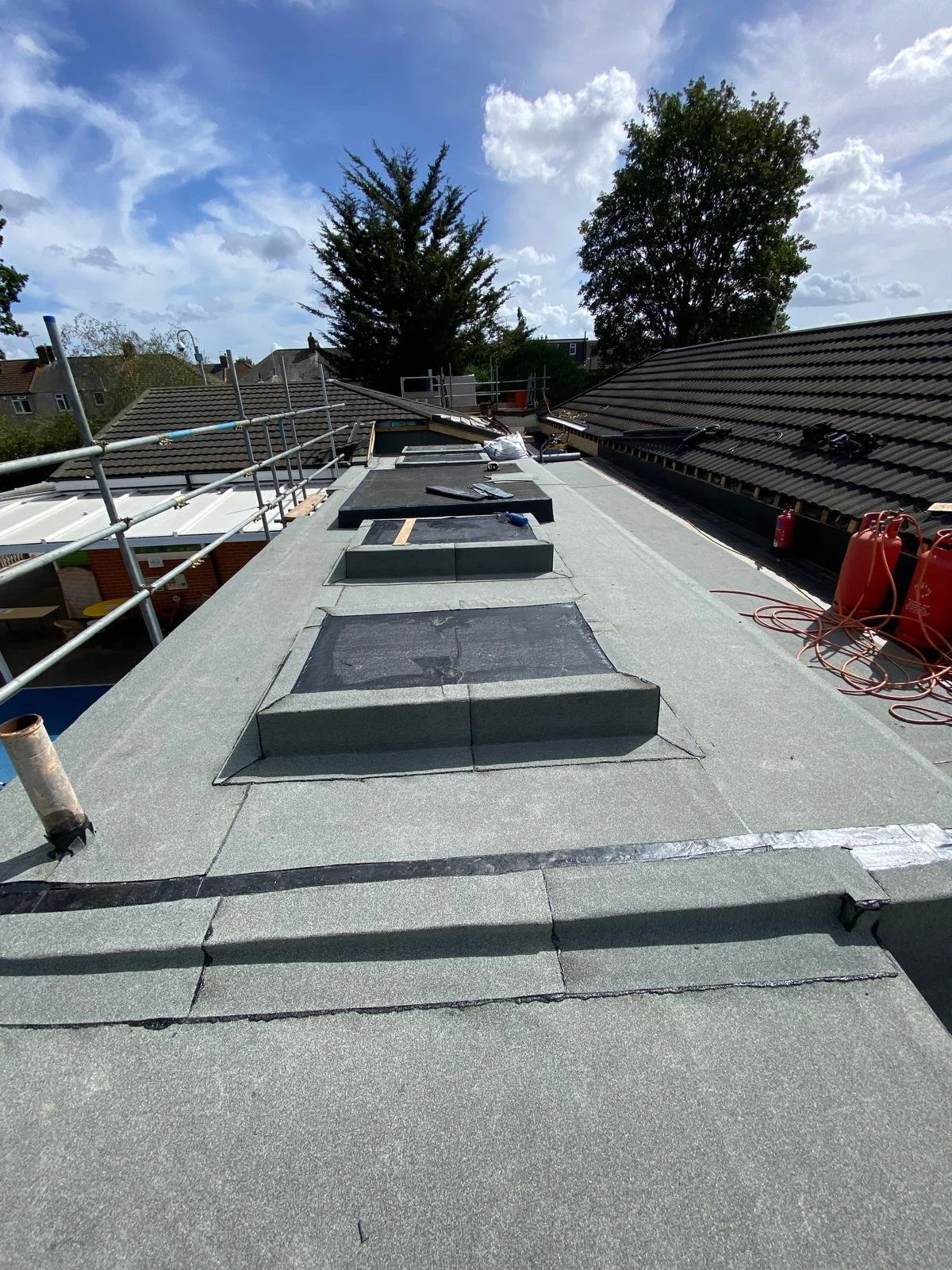 Roof with three skylights being installed, construction tools and supplies visible, on a sunny day with scattered clouds.
