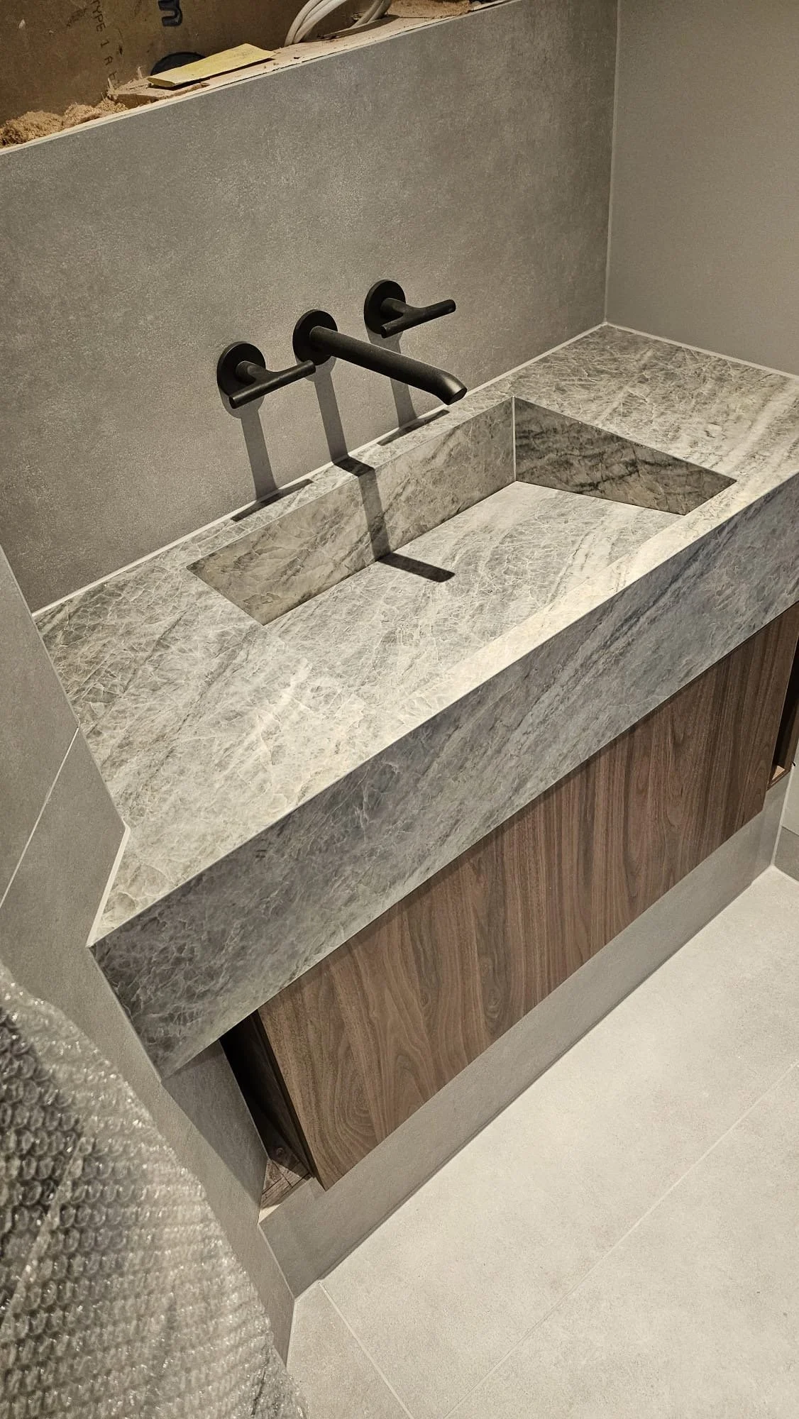 A bathroom vanity with a marble countertop and a rectangular undermount sink. A wall-mounted black faucet is installed above the sink. The wall behind the vanity is unfinished, exposing drywall and construction materials.
