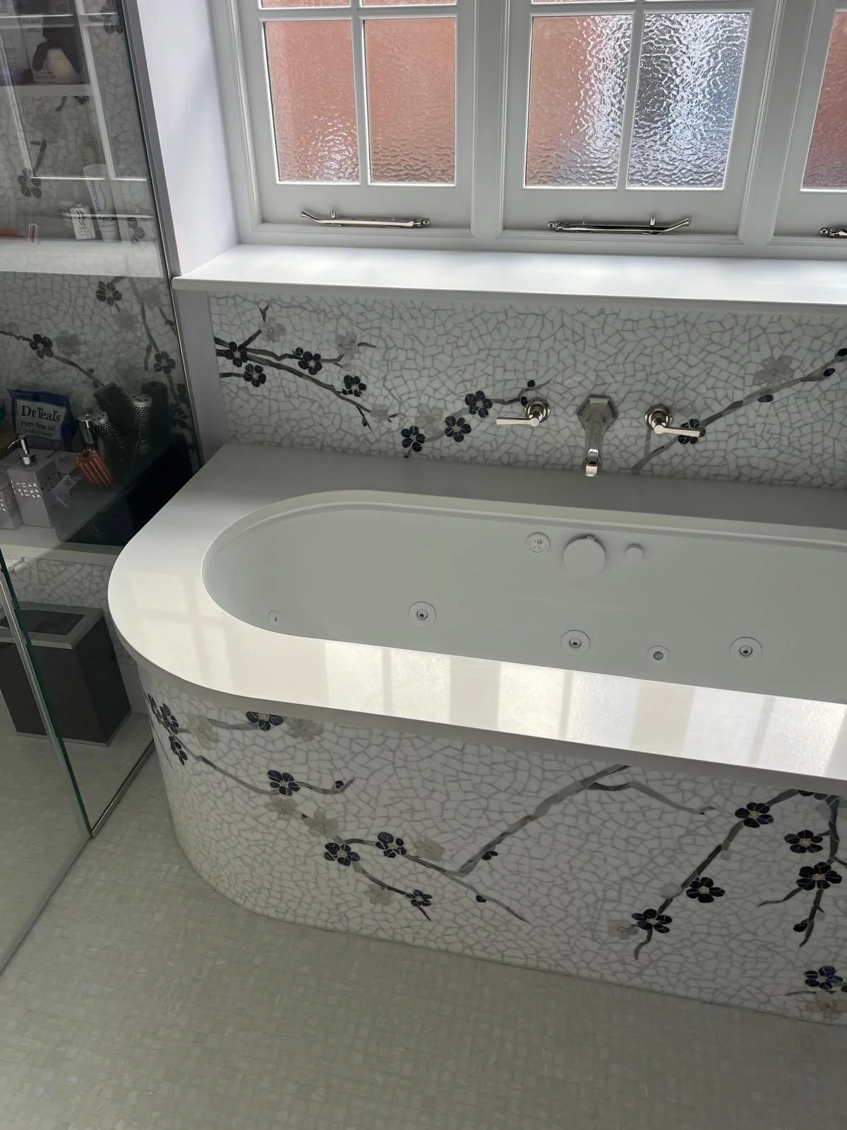 Bathroom with a mosaic tub, floral design on the tiles, and a window above the tub.