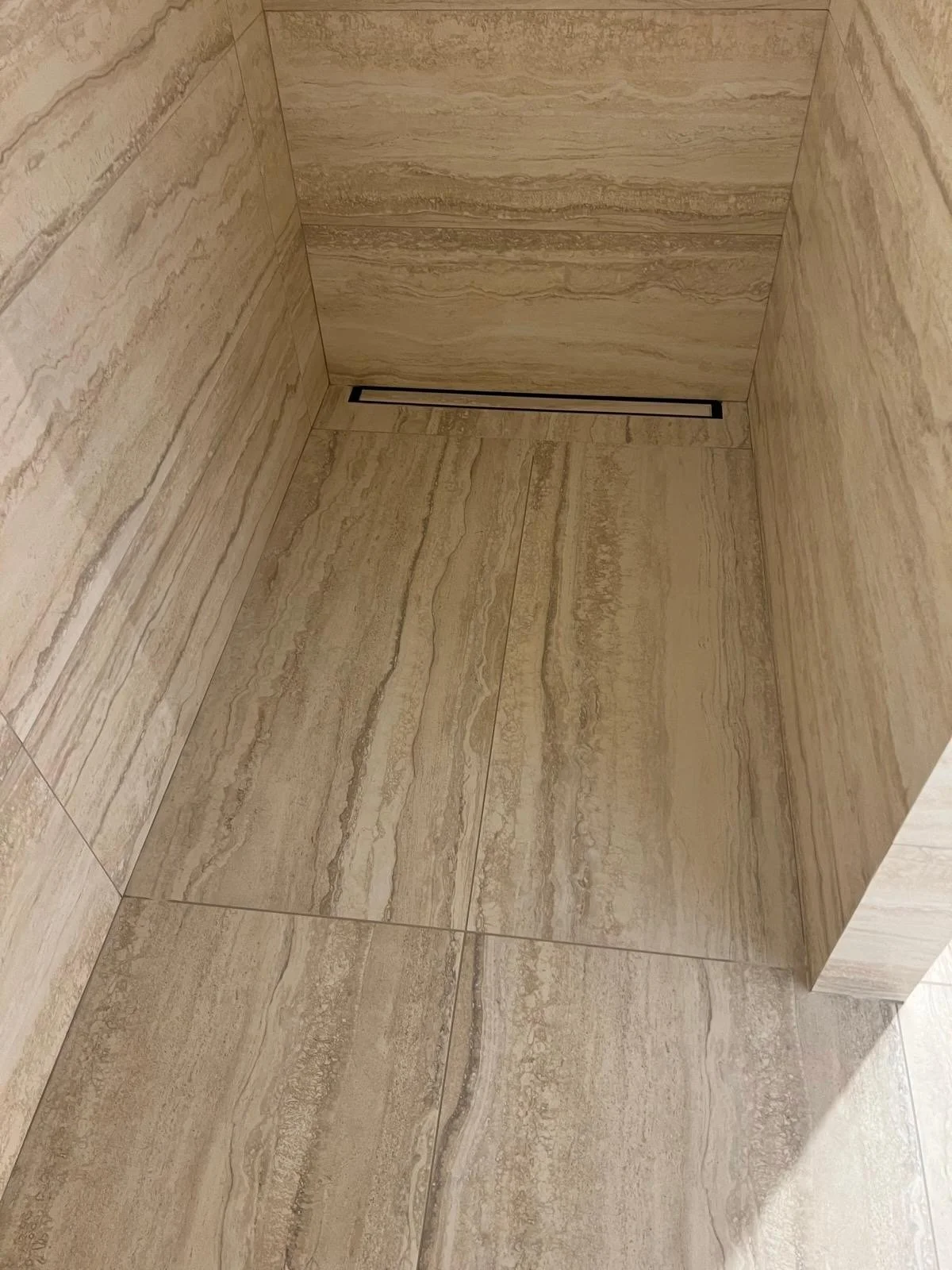 A view of a corner of a tiled room with beige and brown marble-like tiles on the walls and floor, featuring a linear floor drain near the corner.