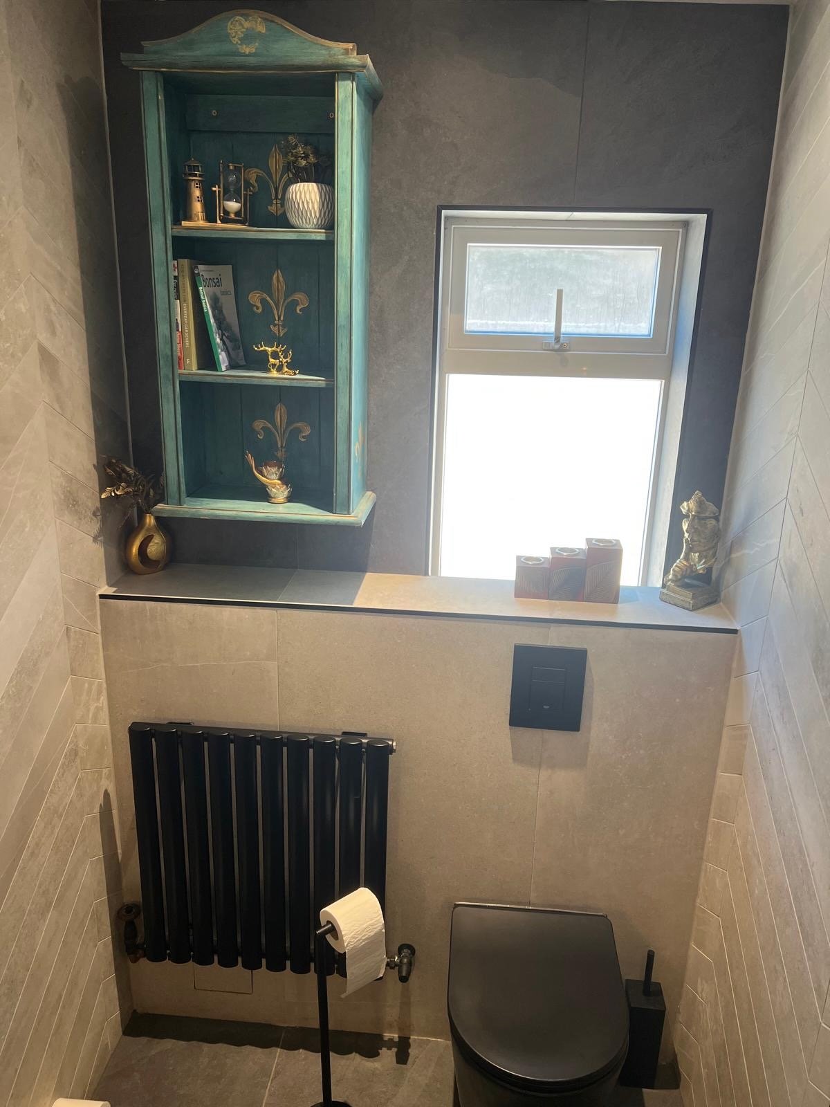 A bathroom with beige tile walls, a black heated towel radiator, a toilet, and a small window. Decorative items such as a golden hand sculpture, a golden deer figurine, and a multicolored square boxes are on the window sill. A blue wall-mounted shelf