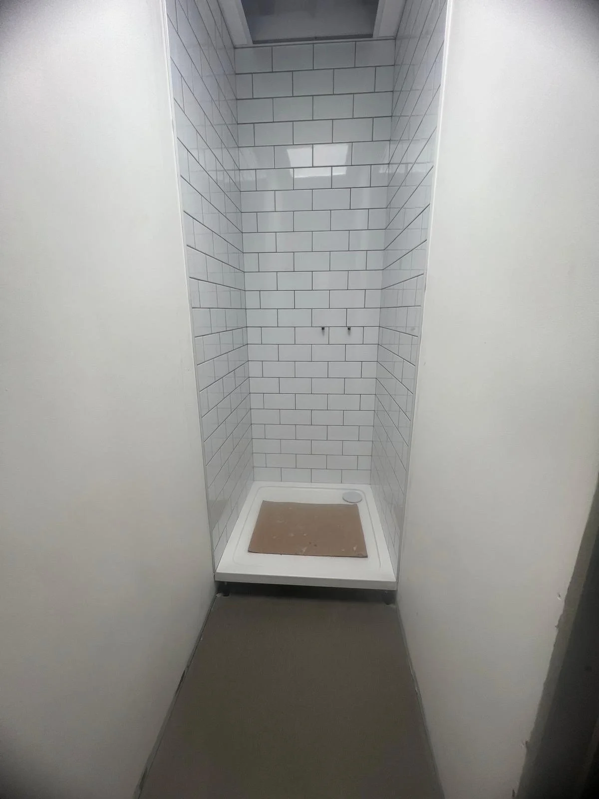 Empty shower stall with white subway tile walls, a small drain, and a rectangular beige mat on the floor.