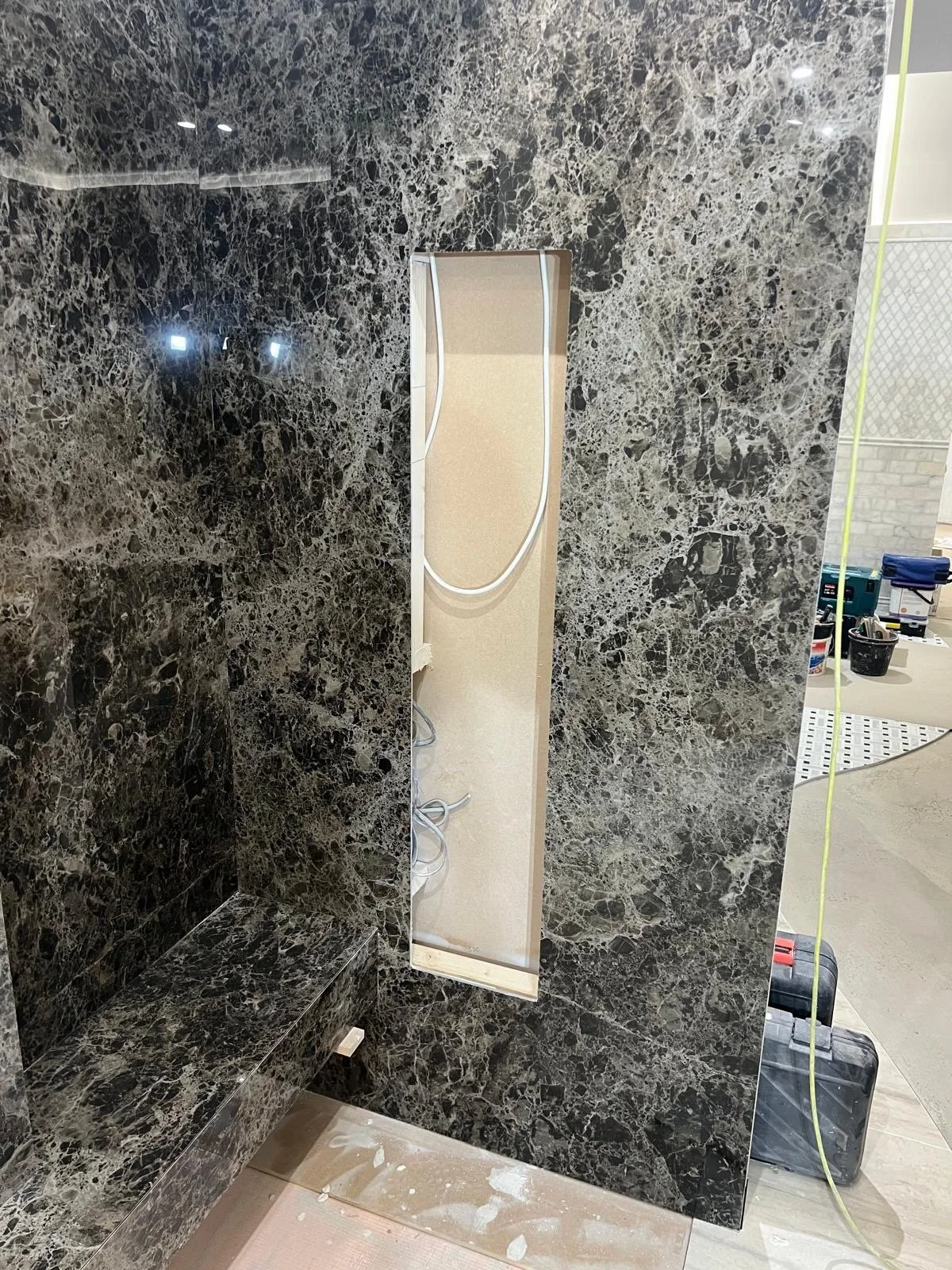 A bathroom wall with black marble tiles and an empty rectangular mirror frame. Construction tools and supplies are visible in the background.