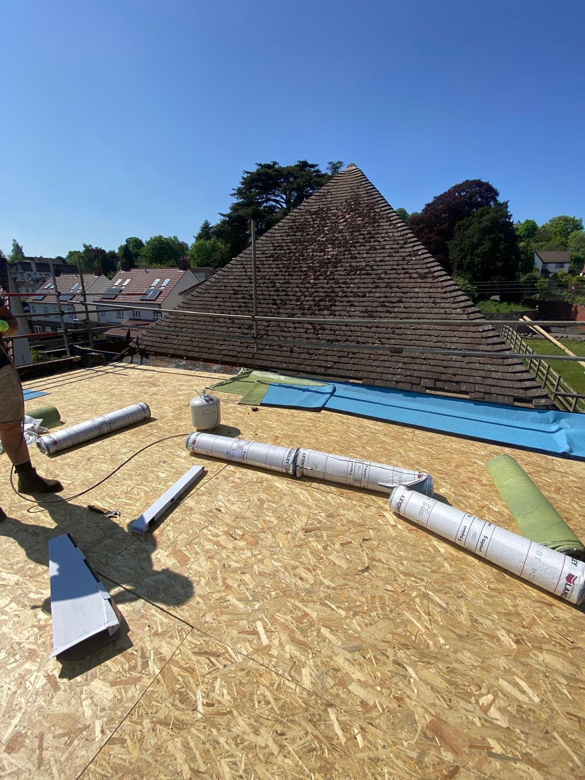Roof under construction with rolled roofing materials and blue and green tarps, a partially visible scaffold, and a background of trees and neighboring rooftops on a sunny day.