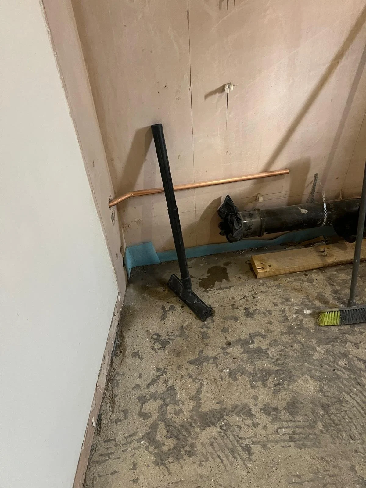 Interior of a construction or renovation site with plumbing and tools, including a black pipe, copper pipe, broom, and a piece of wood on the floor.